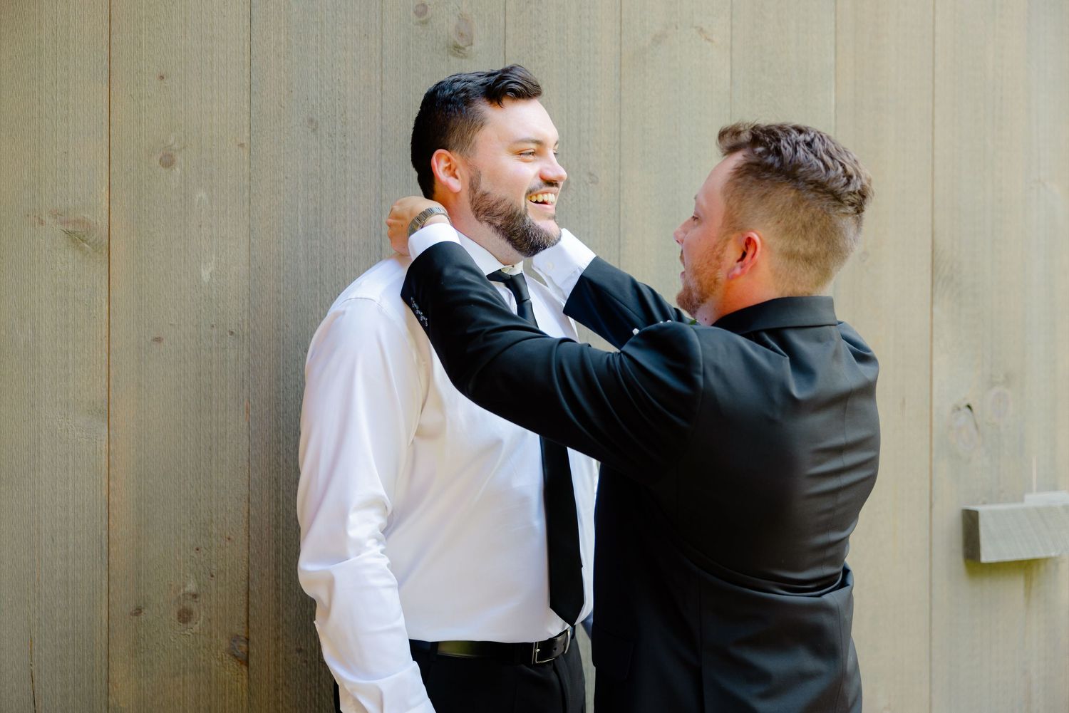 Two men in formal attire share a moment while fixing bow tie before wedding ceremony.