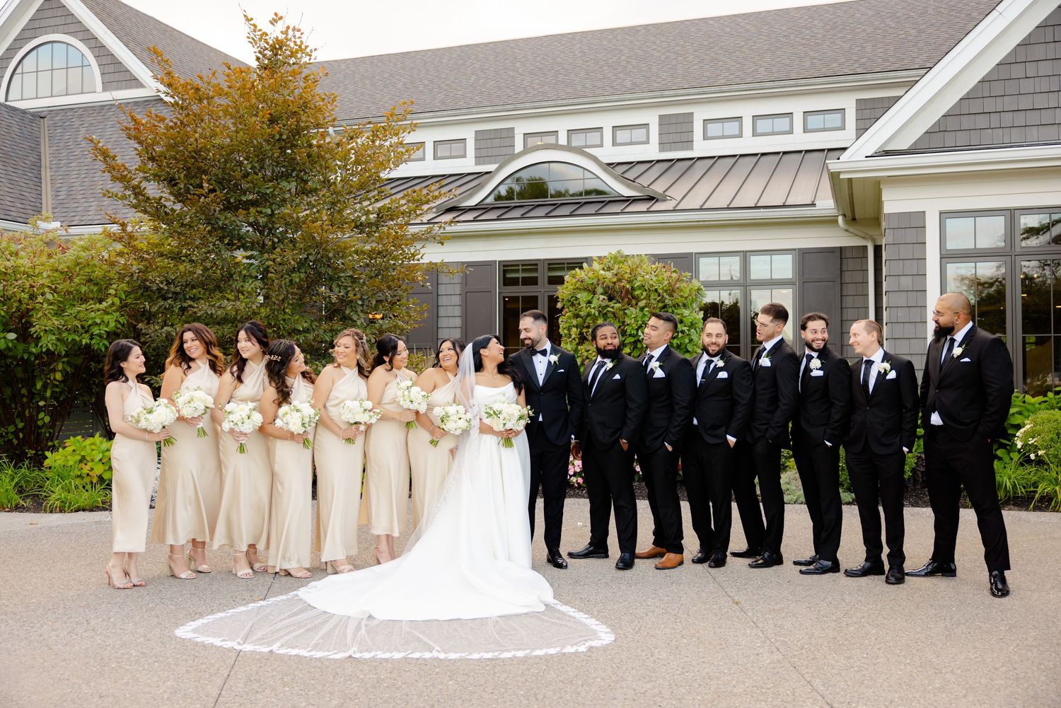 Formal wedding party photo with attendants in cream and black attire outside a coastal venue.