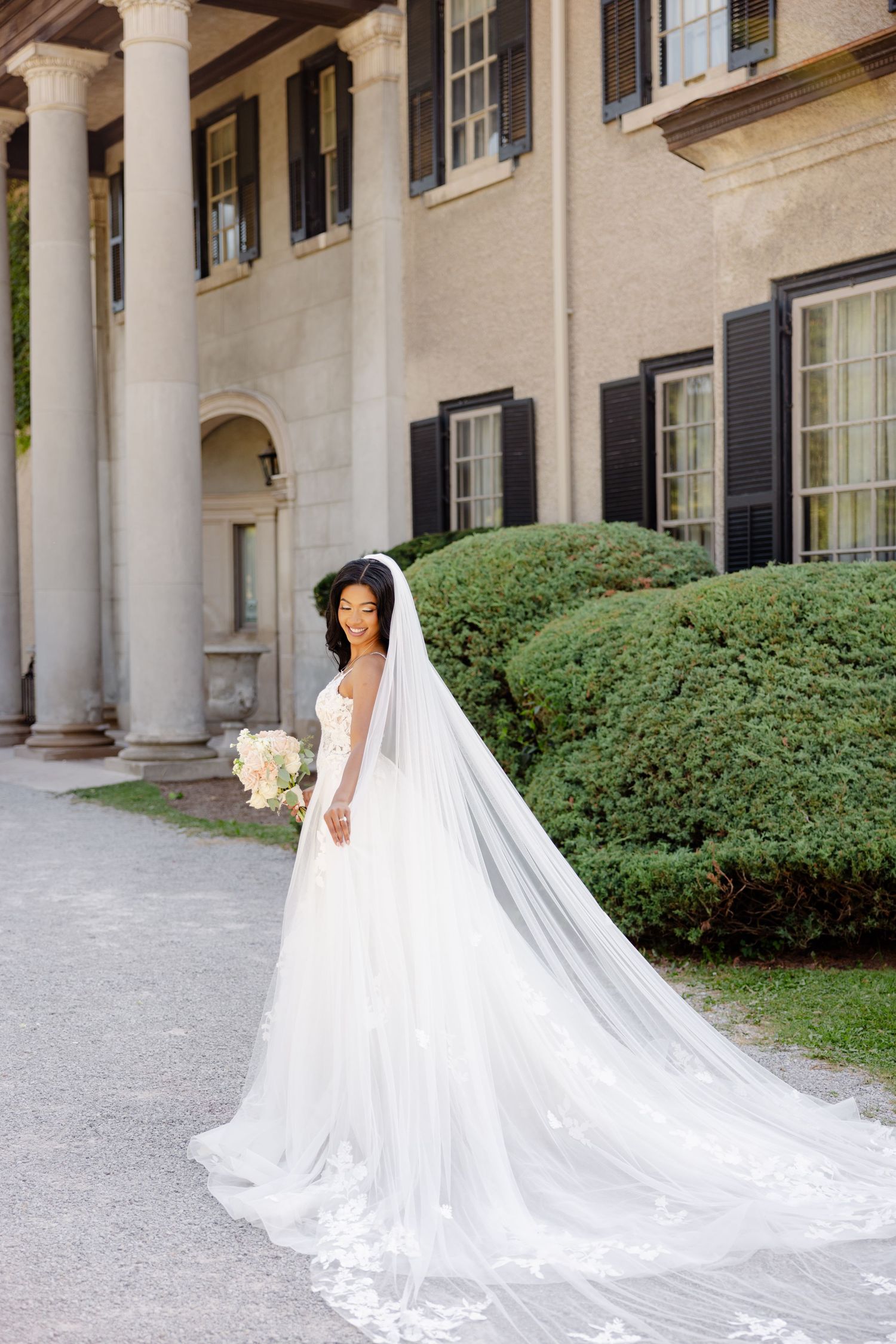 Bride in white gown and cathedral veil poses beside columned building entrance.