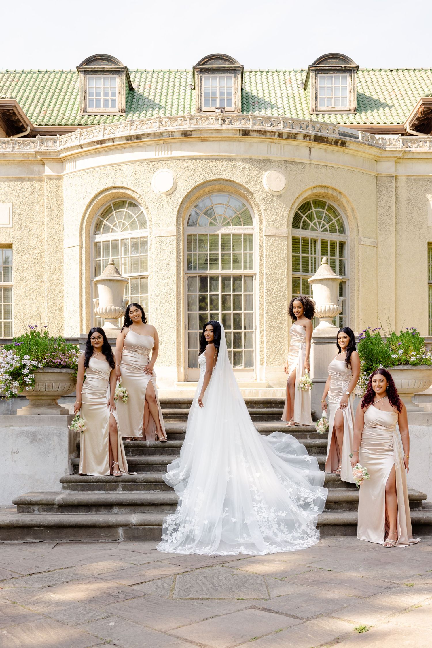 A bridal party poses on grand stone steps wearing champagne dresses in front of a historic mansion with arched windows.