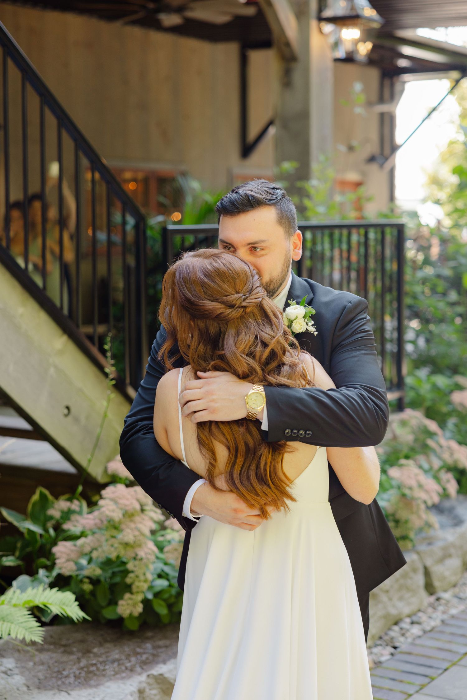 A tender embrace between two people in wedding attire on a stone pathway surrounded by lush greenery.