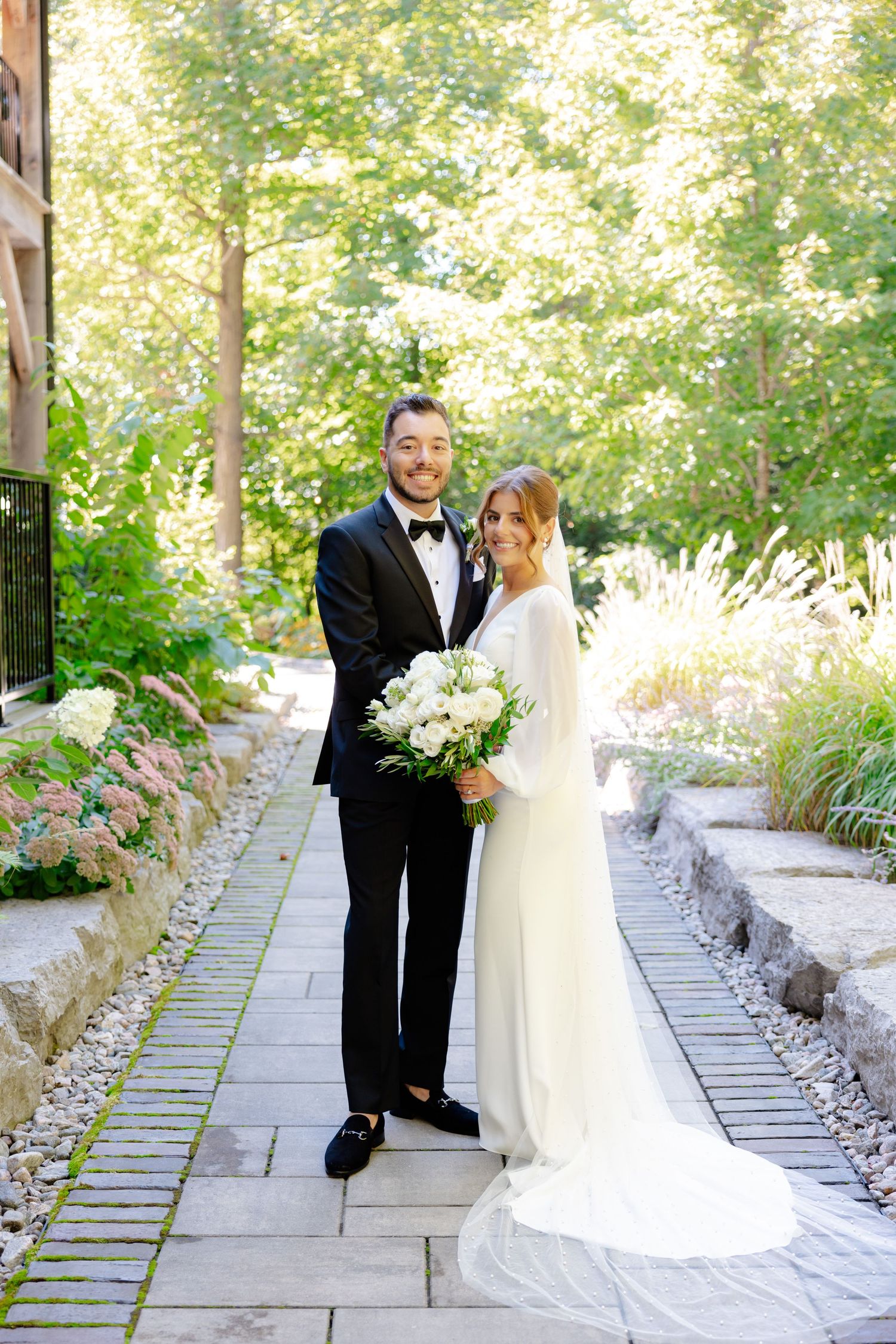 Elegant wedding couple stands on stone path surrounded by gardens holding white flower bouquet.