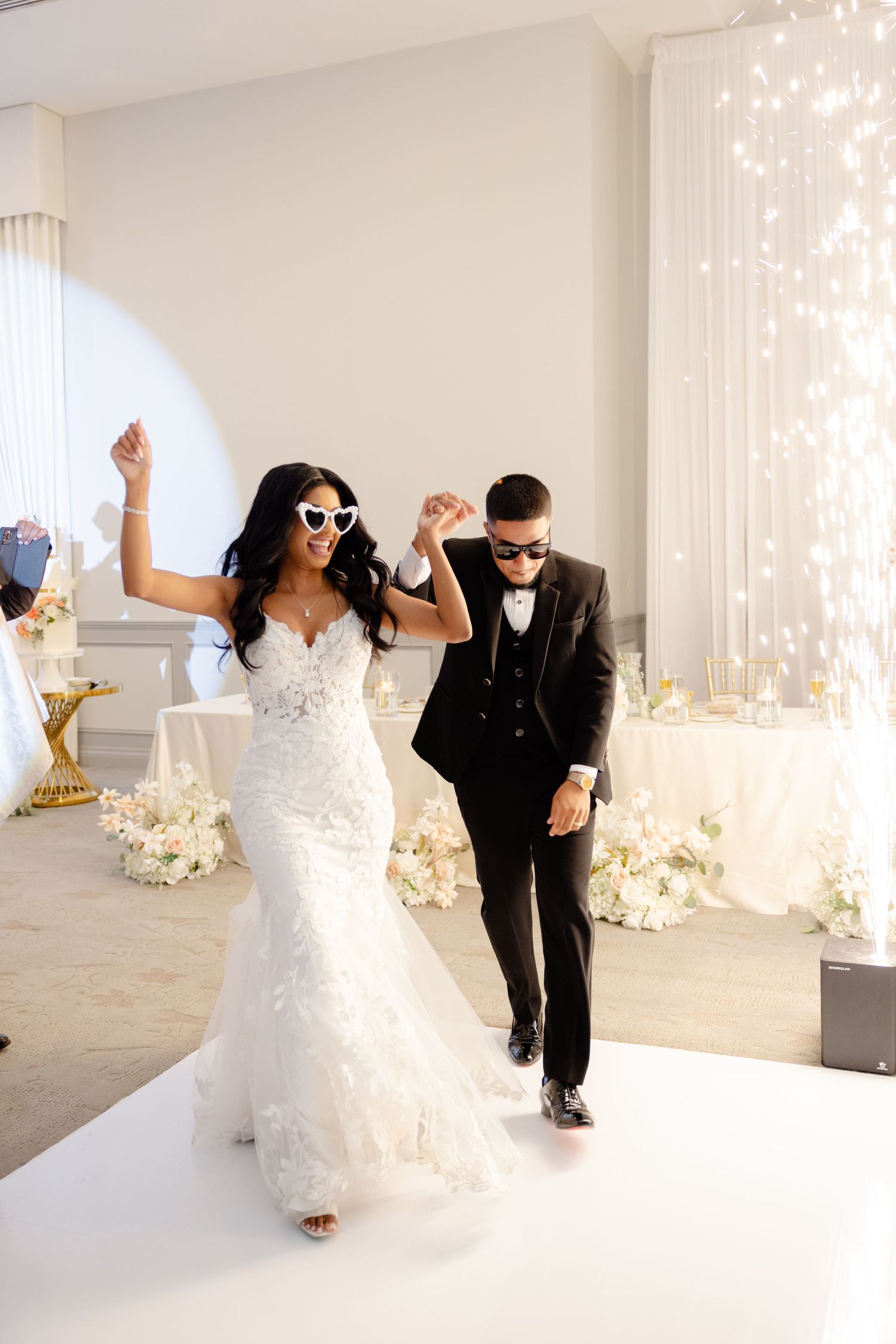 Newlyweds wearing sunglasses dance together on a white platform with sparkly backdrop and floral decorations.