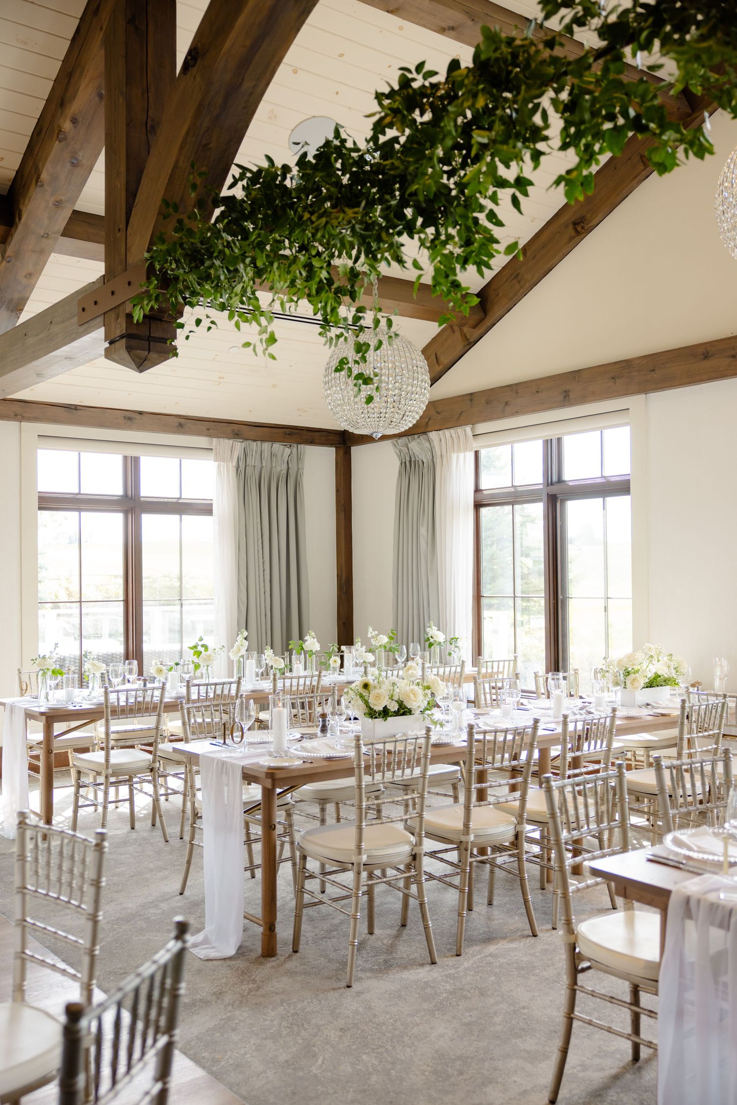 Light-filled wedding reception space with exposed beams, white decor, and trailing greenery installations.