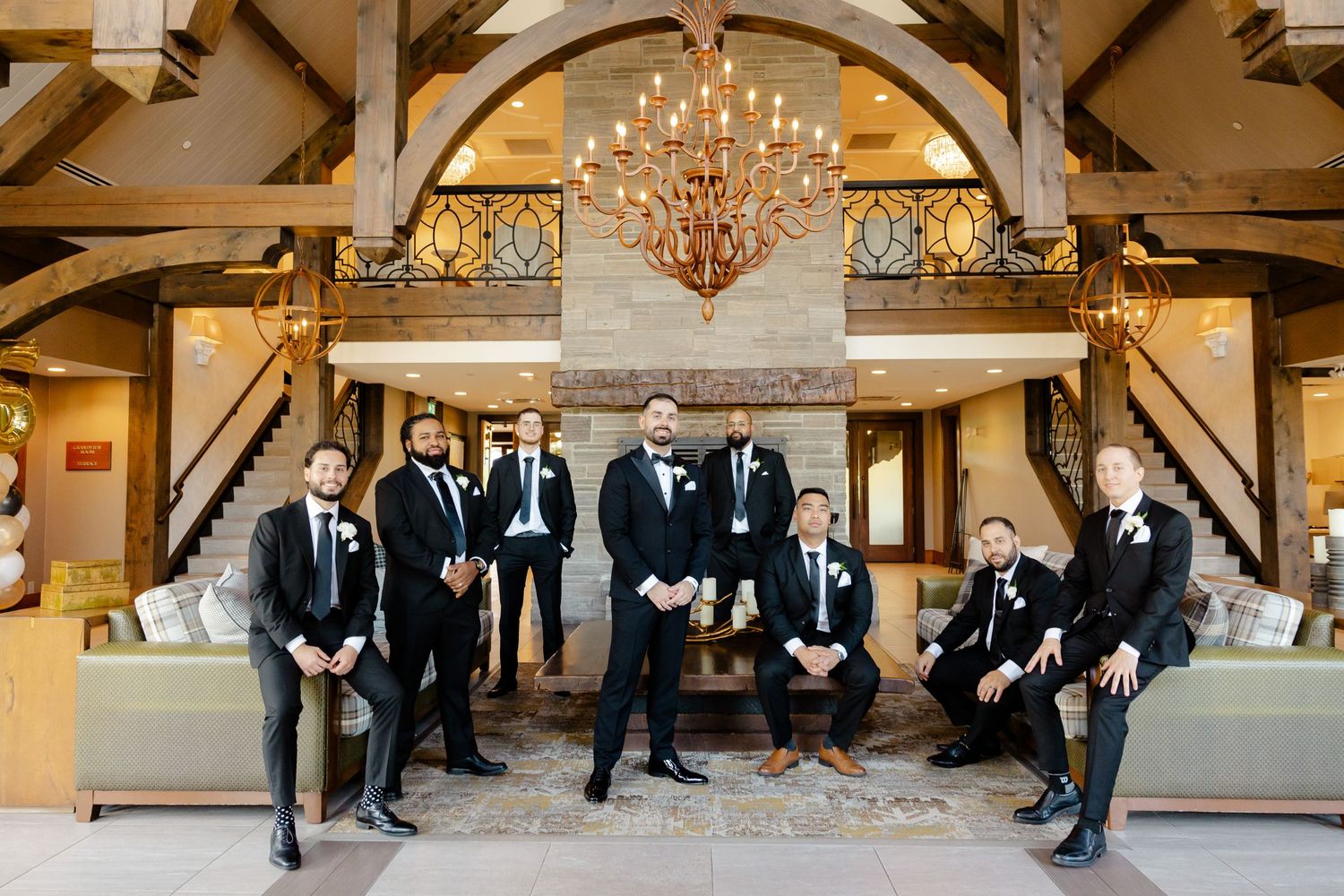 Wedding party in formal attire poses inside rustic venue with wooden beams and chandelier.