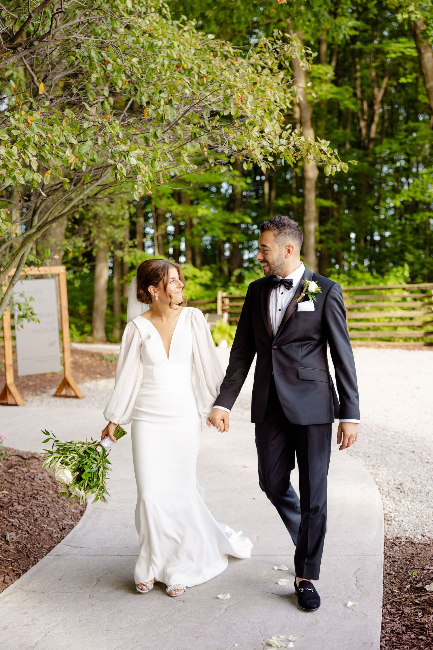 A couple walks hand in hand along a wooded path, with the bride in a white gown and groom in classic tuxedo.
