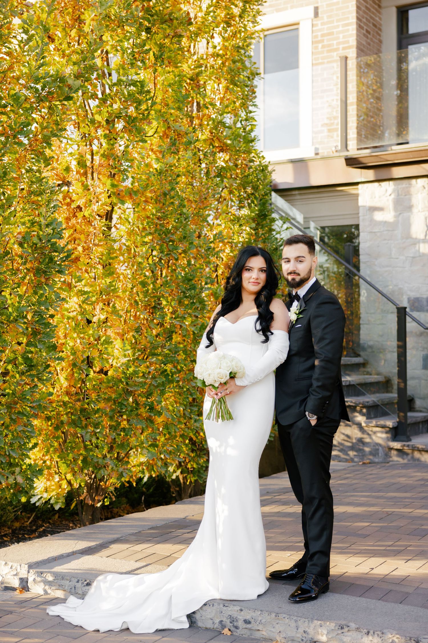 Wedding portrait with fall foliage backdrop, featuring bride in white gown and groom in classic black suit.