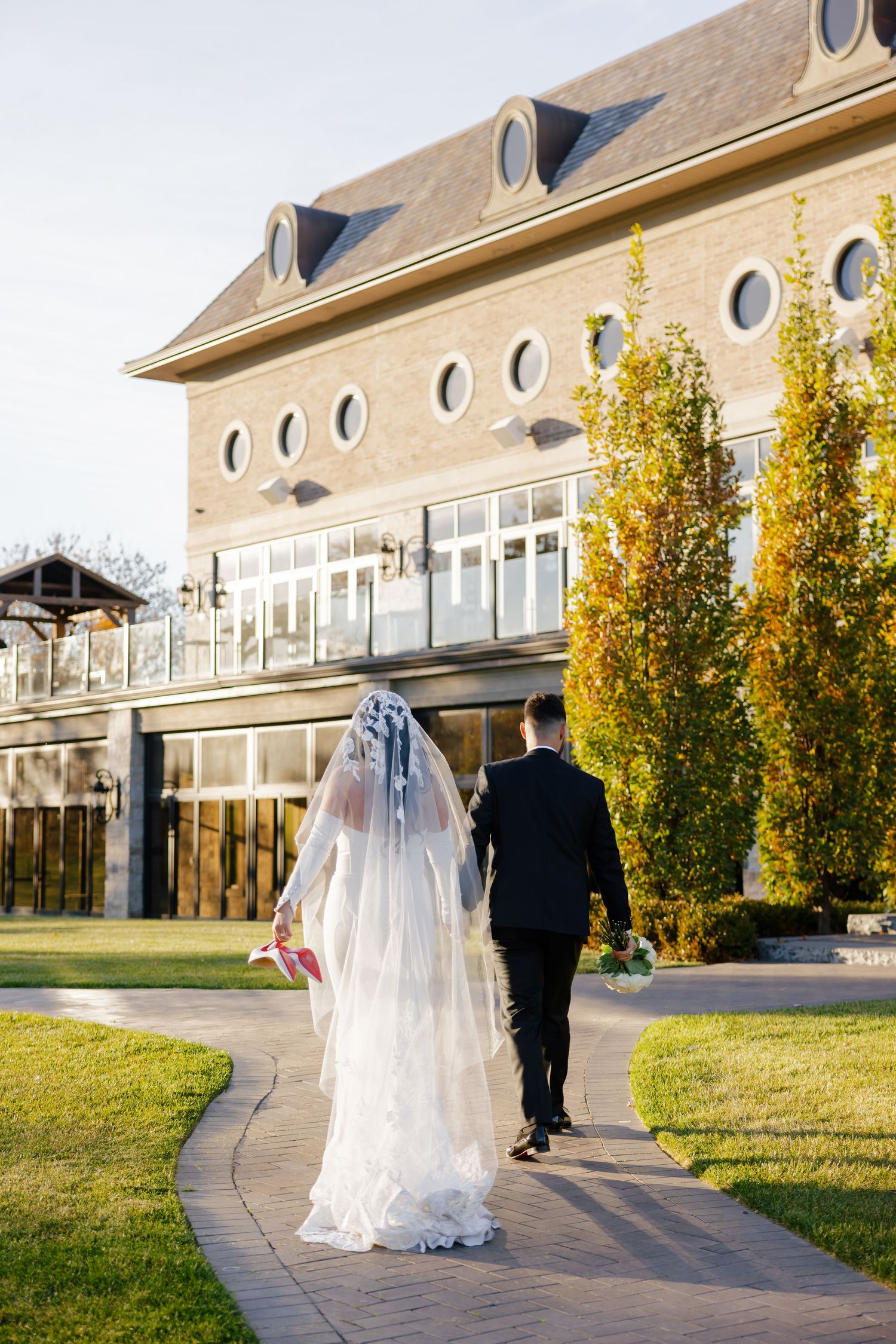 Wedding couple walks hand in hand on garden path by elegant building with circular windows in autumn.