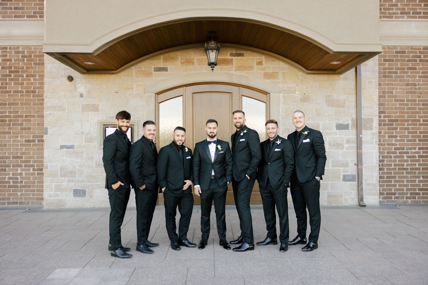 Groomsmen in black suits pose for a formal photo under an arched entranceway.