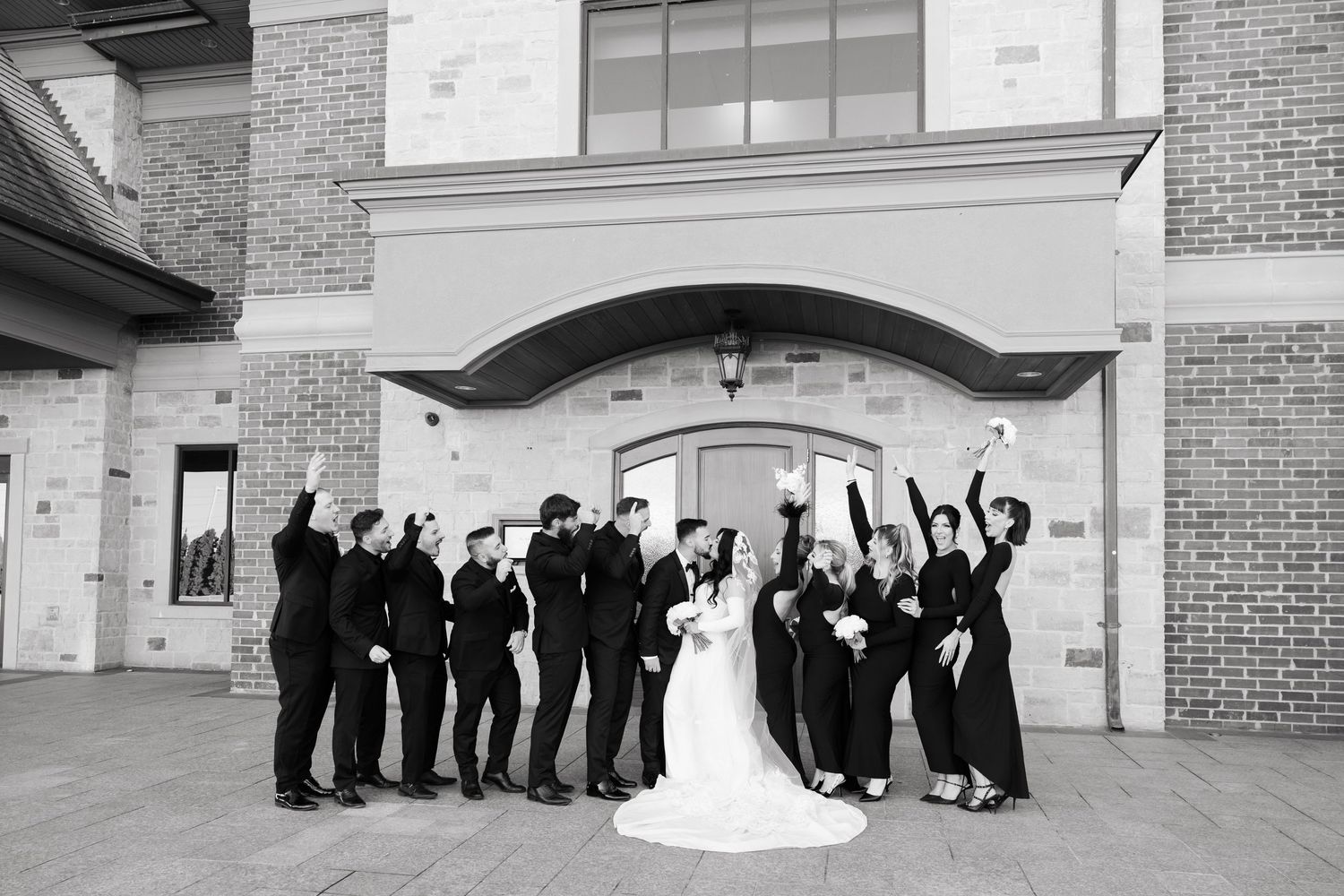 Black and white photo of wedding party celebrating with energetic poses outside brick building.