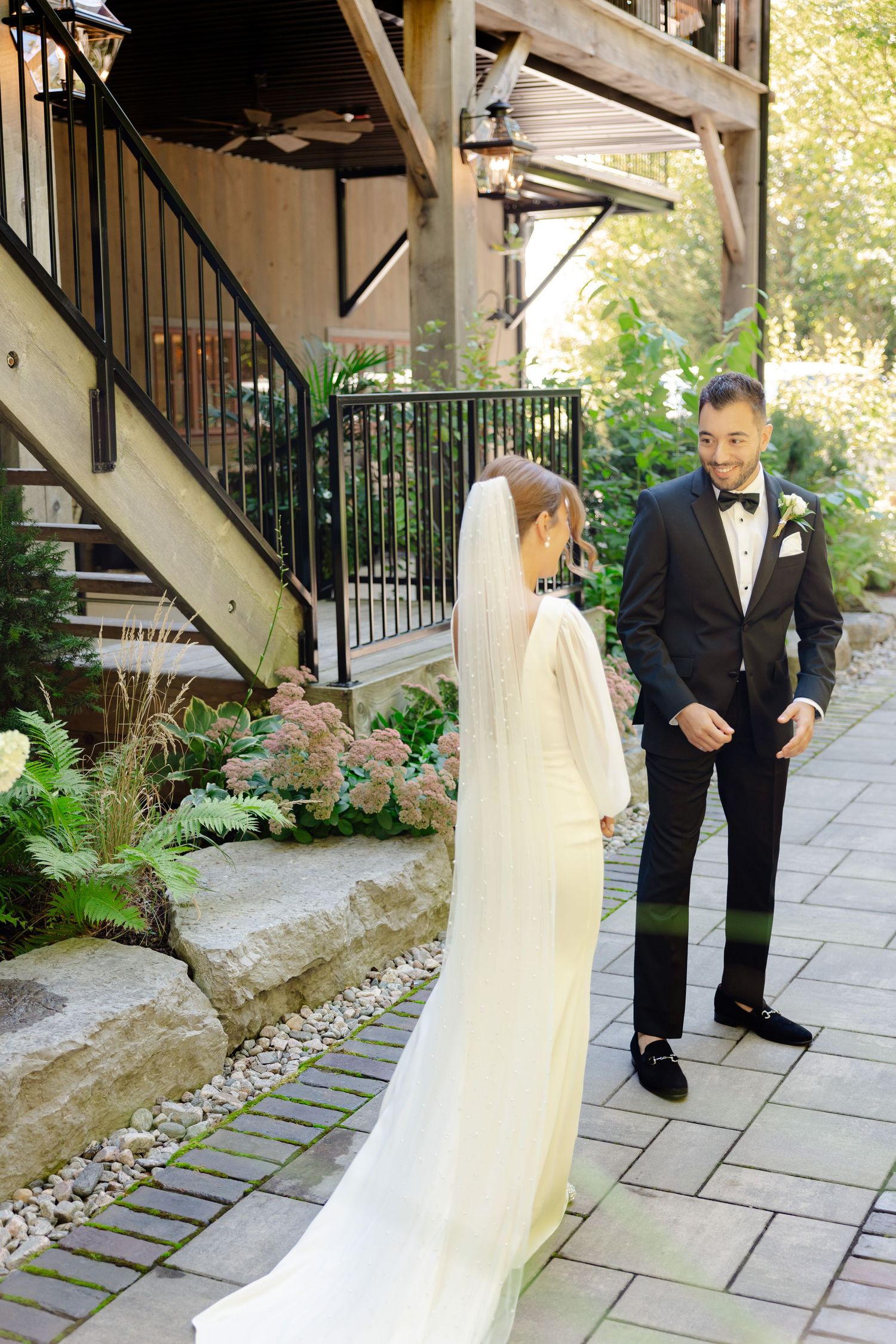 A couple shares their first look moment on a stone-paved garden path with lush greenery and wooden stairs.