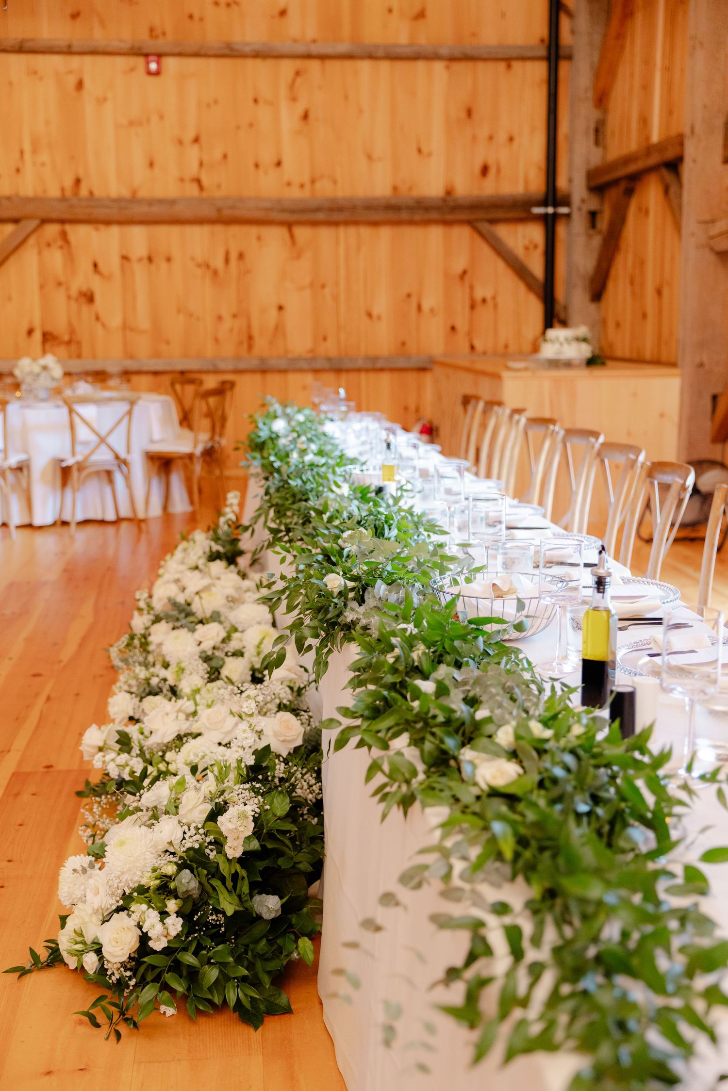 Long banquet table with lush white floral arrangements and greenery garlands in a rustic barn setting.