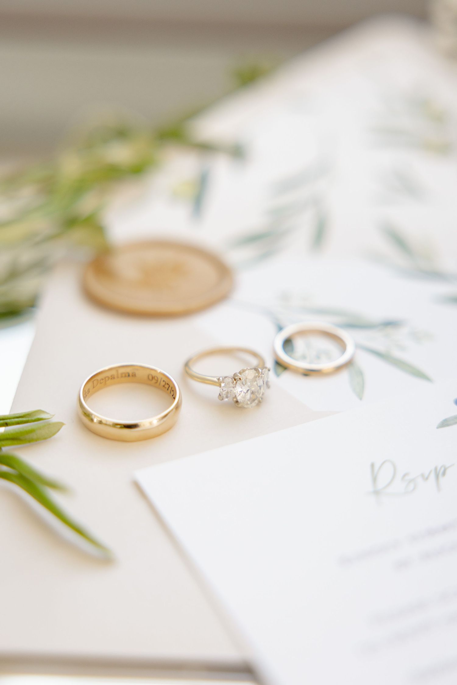 Wedding rings and engagement ring arranged on white stationery with greenery accents.