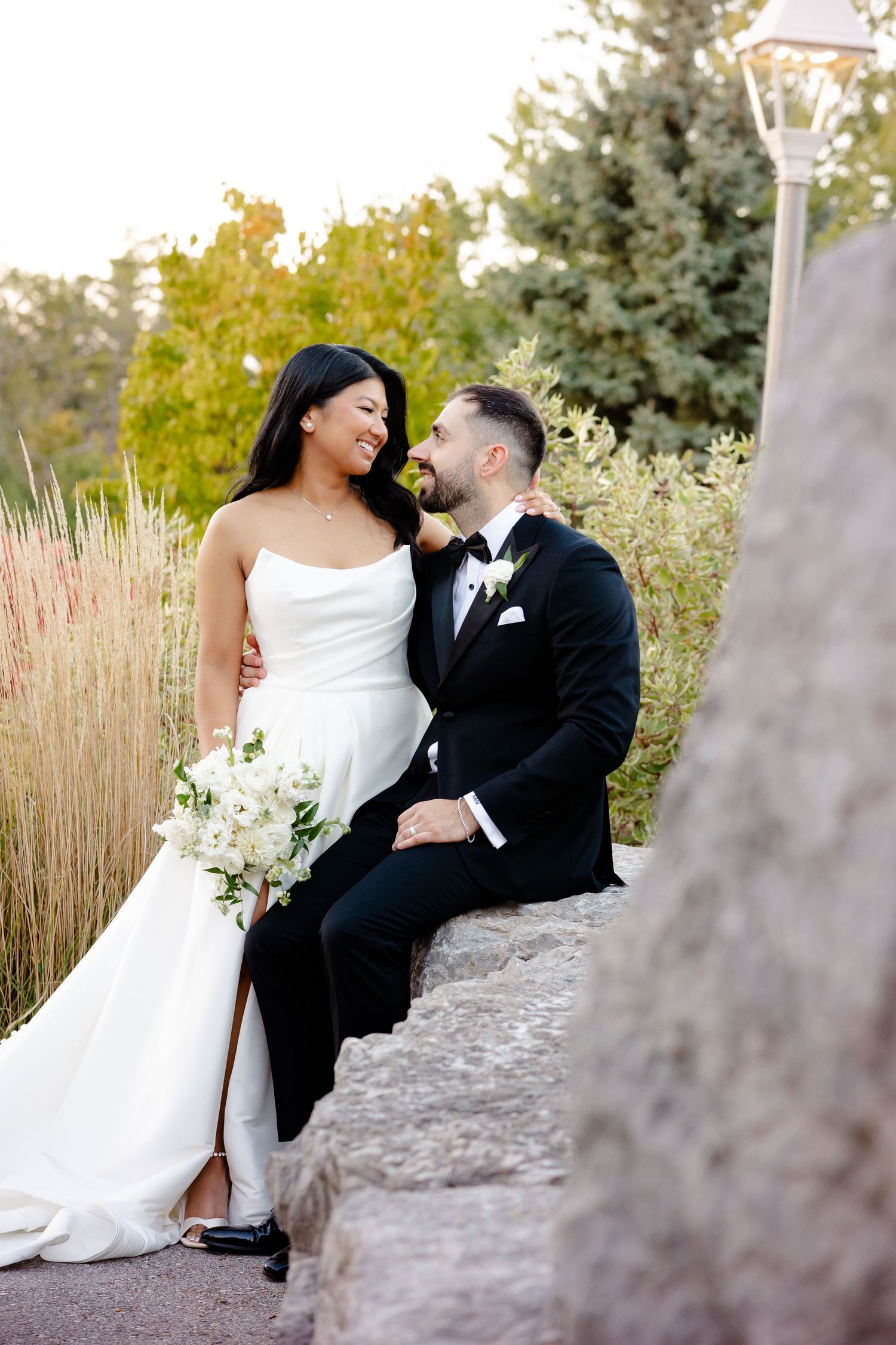 A joyful wedding couple shares a laughing moment while sitting on stone steps amid ornamental grasses.