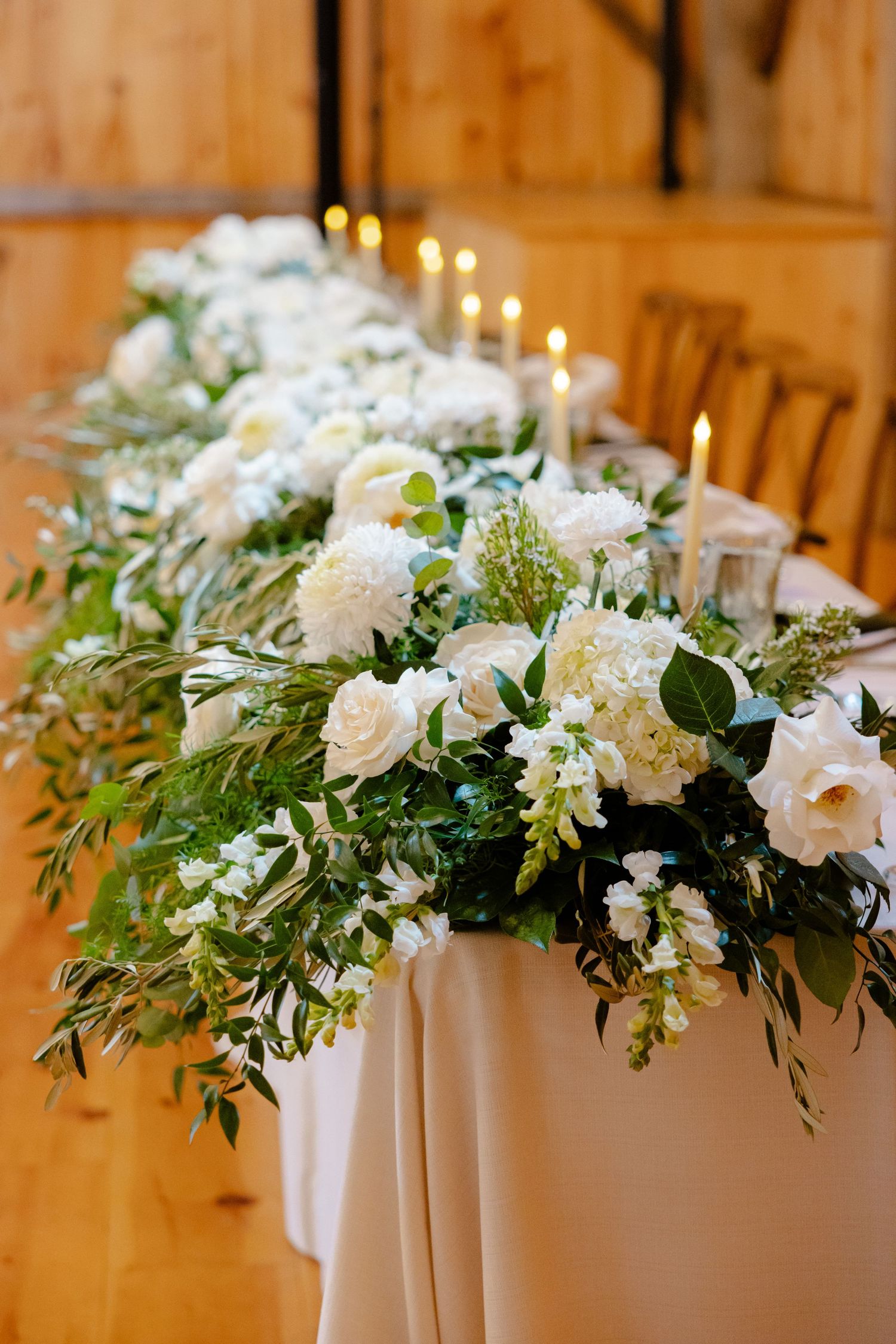 A lush white and green floral table arrangement with roses and mixed foliage on a white tablecloth.
