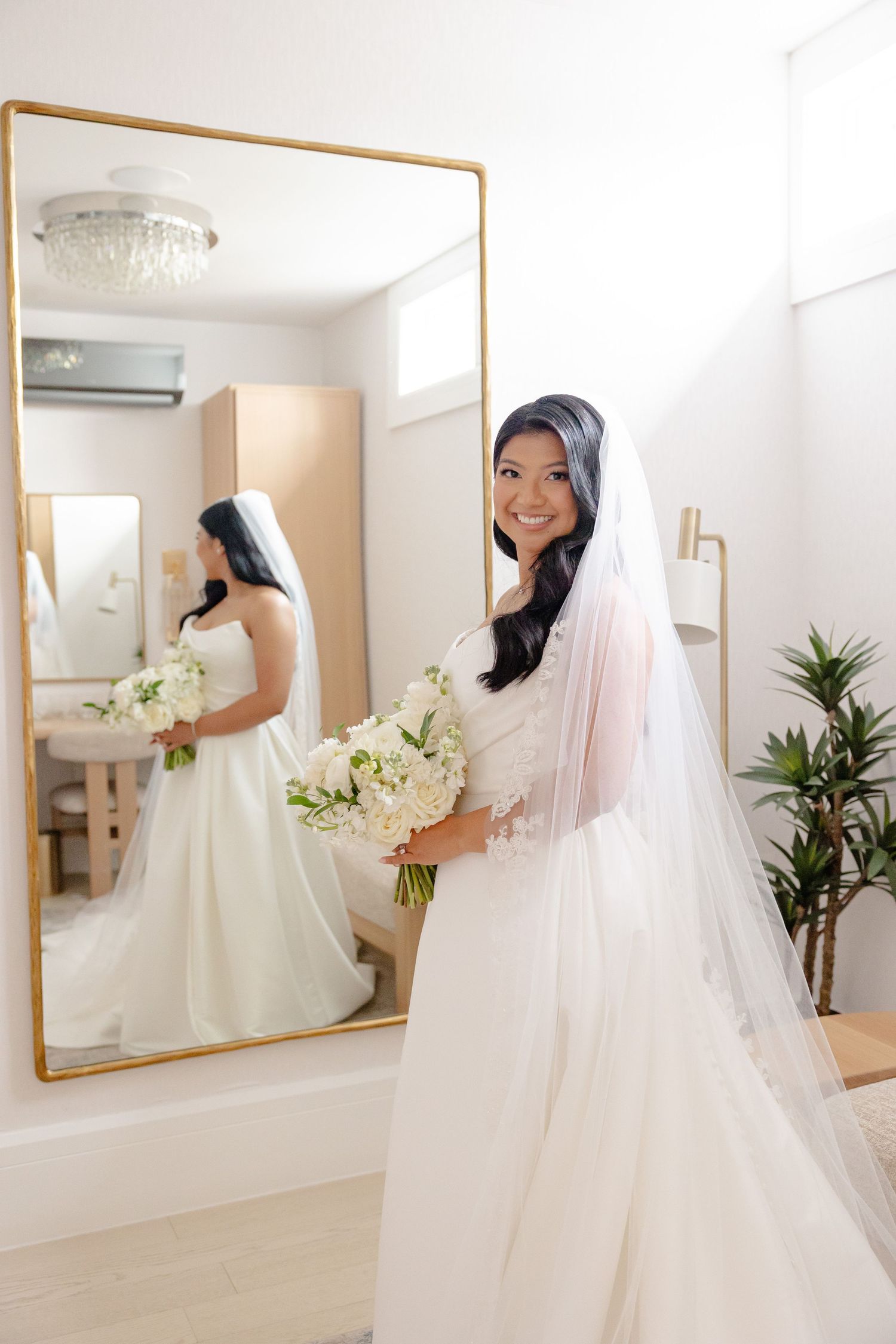 Bride poses with bouquet beside mirror reflection in bright wedding prep room.