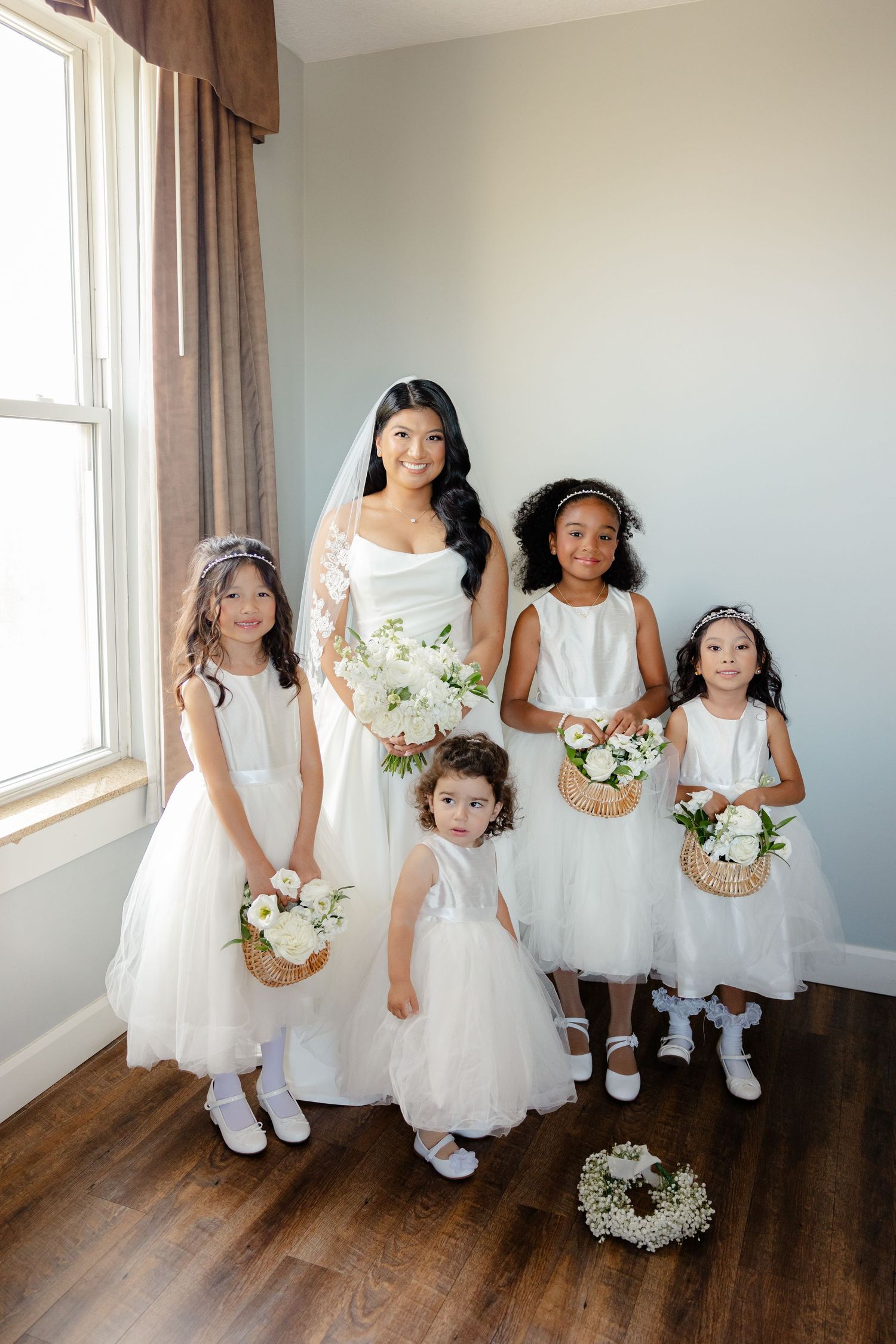 Flower girls in white dresses carry floral baskets while posing with the bride by a window.