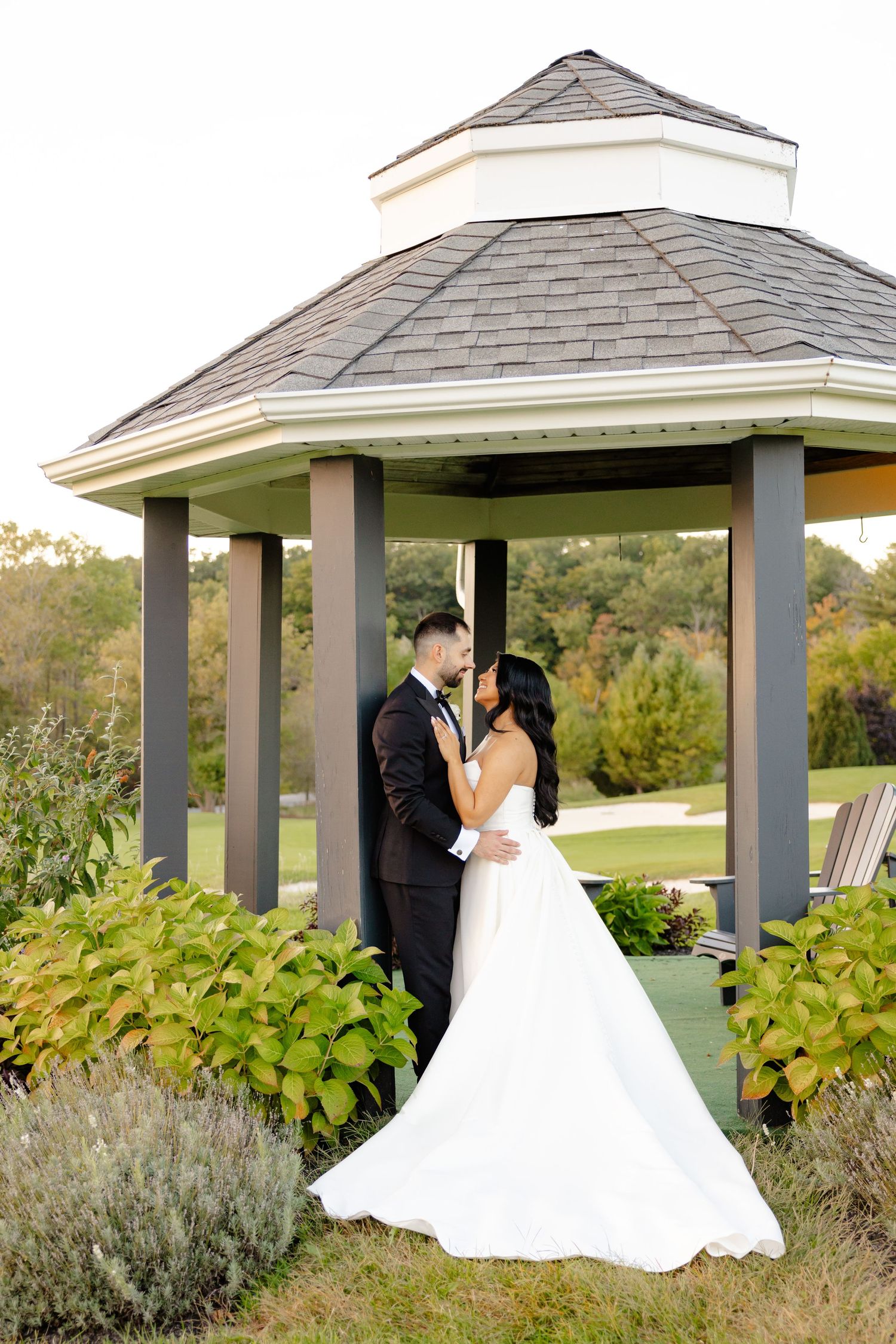 Romantic wedding portrait of couple under garden gazebo surrounded by lush landscaping.