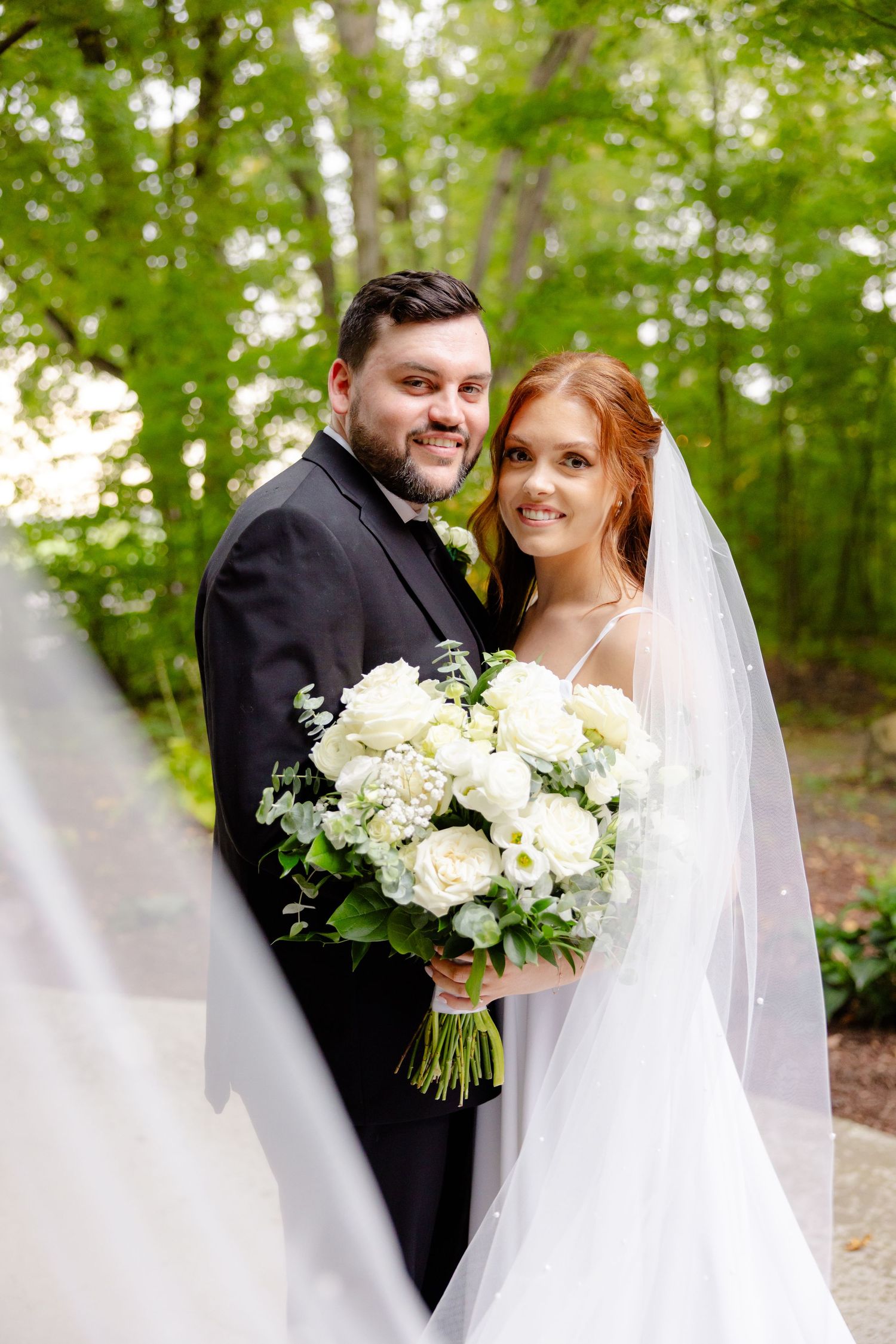 Joyful wedding portrait in a wooded setting with elegant white bouquet and flowing veil.