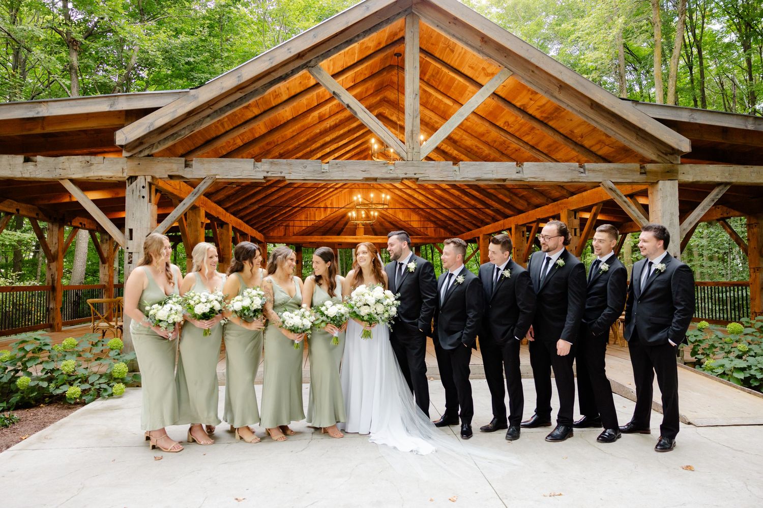 Full wedding party in formal attire poses under rustic wooden pavilion with string lights.