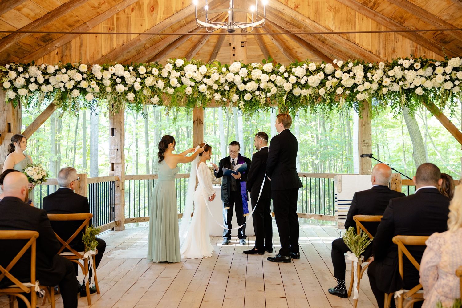Wedding ceremony taking place in a rustic wooden gazebo decorated with white floral arrangements and greenery.