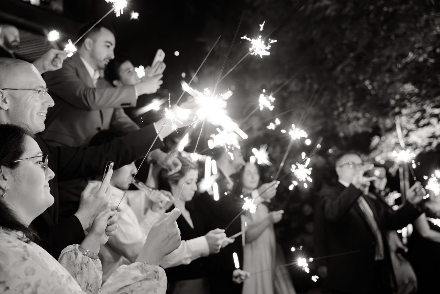 Wedding guests hold sparklers creating a magical black and white nighttime atmosphere.