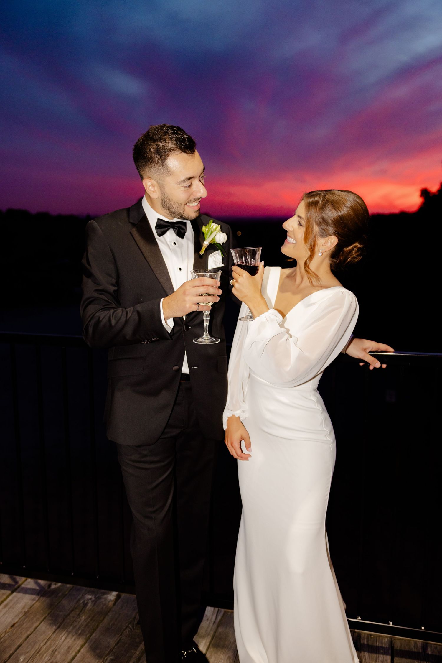 A newlywed couple toasts with champagne glasses against a dramatic purple sunset sky.