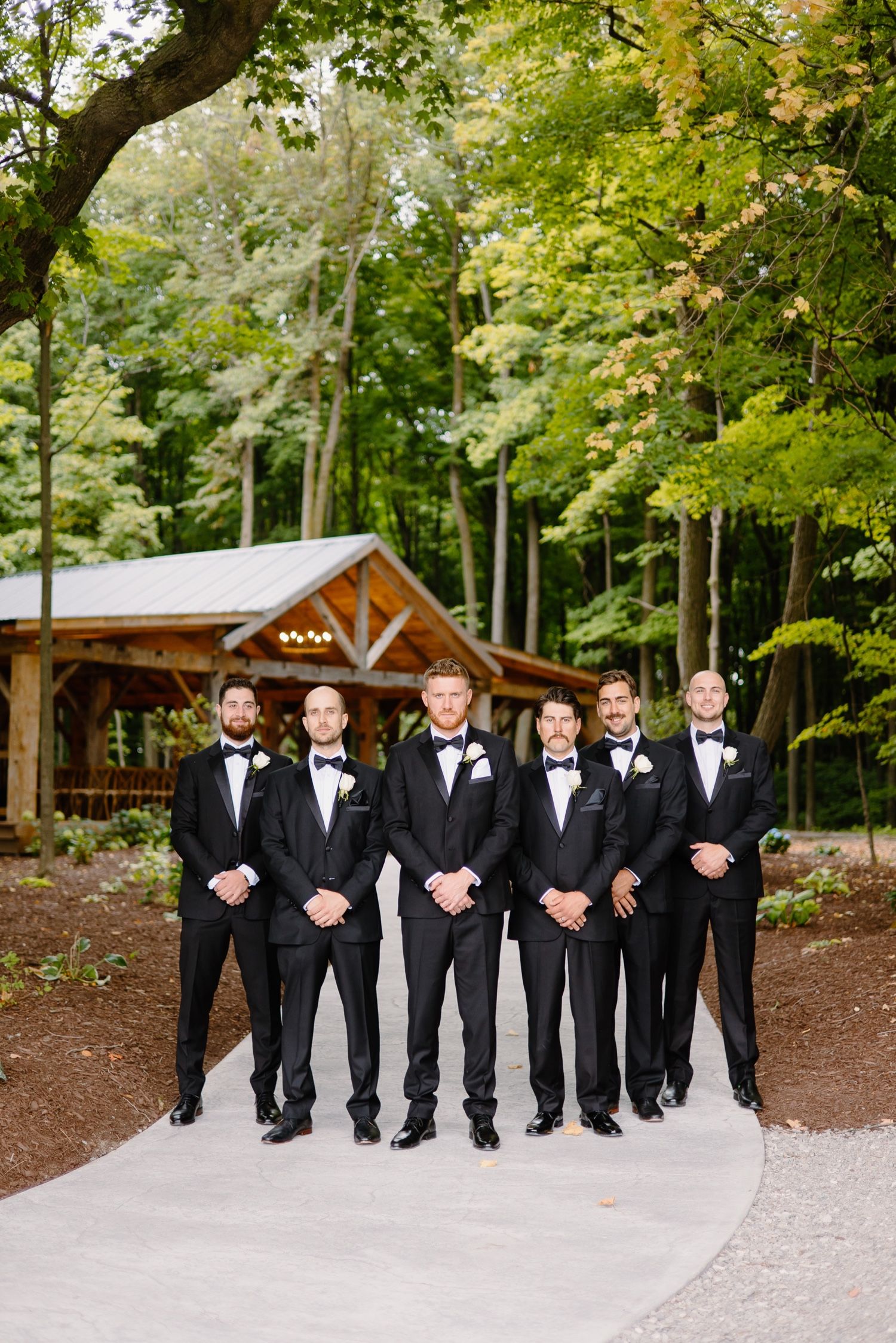 Groomsmen line up in matching tuxedos on pathway with rustic cabin in background.