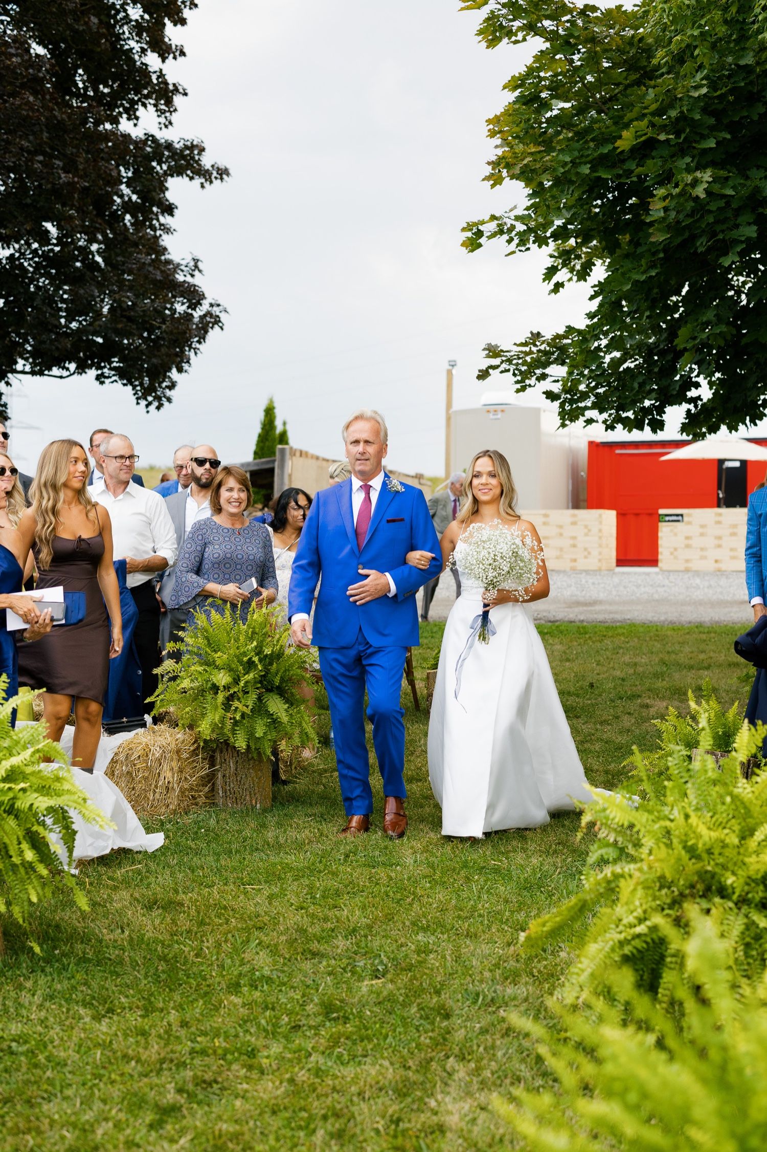 Elegant outdoor wedding processional with attendants walking down aisle lined with ferns.