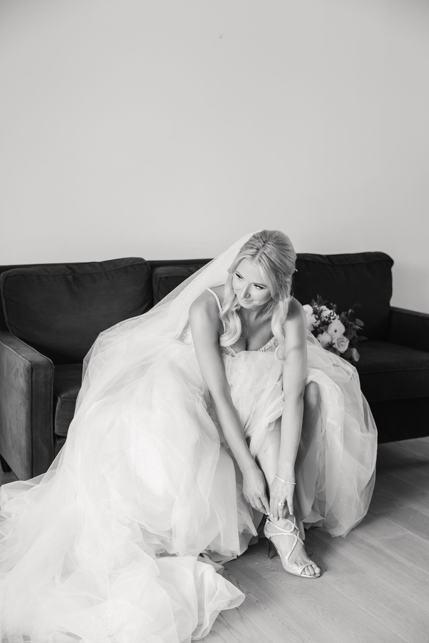 Black and white image of bride fastening her elegant wedding shoes while seated on dark sofa.