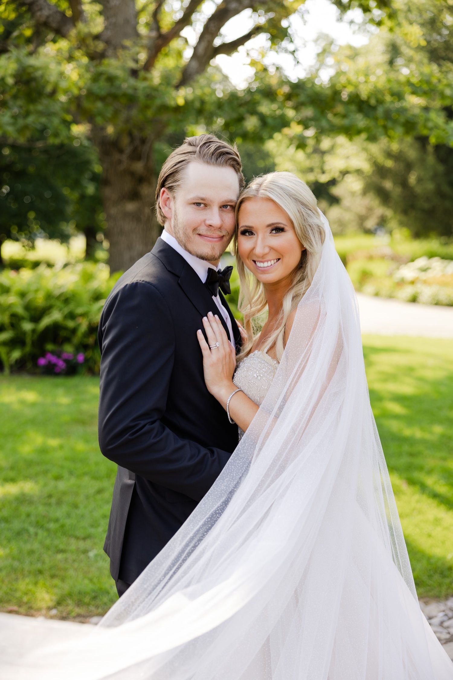 A romantic wedding portrait featuring a couple in formal attire with a flowing veil in a lush garden setting.