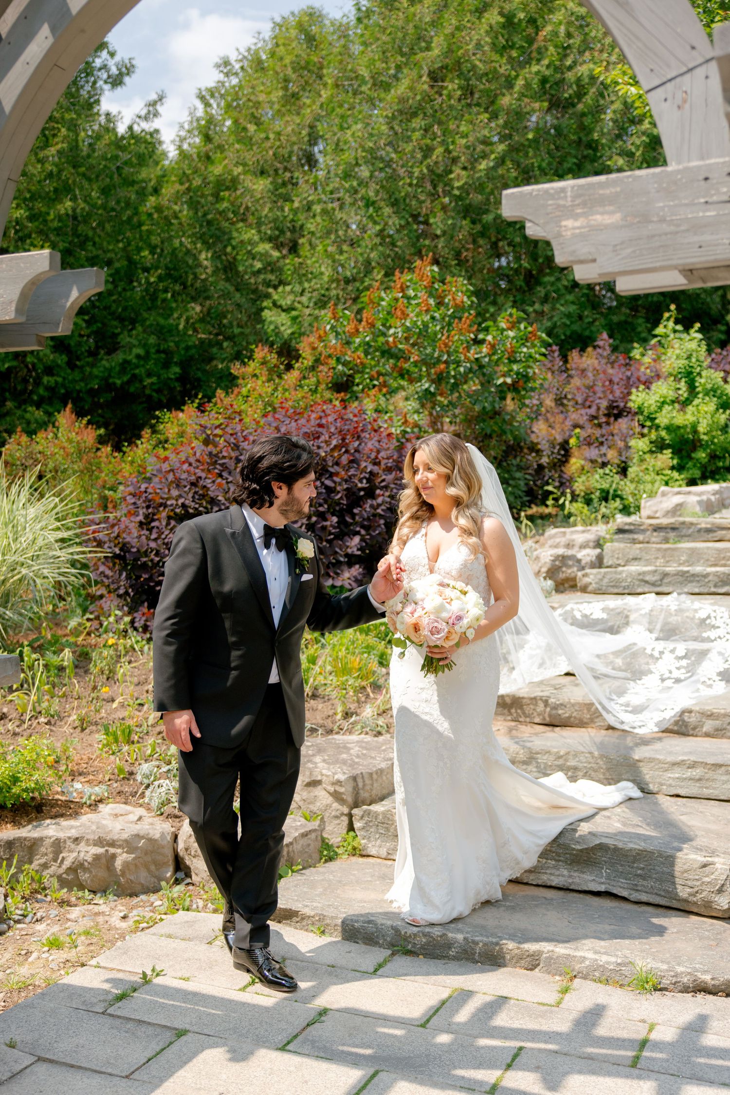 A couple shares a moment on stone garden steps framed by a wooden arch and vibrant landscaping.
