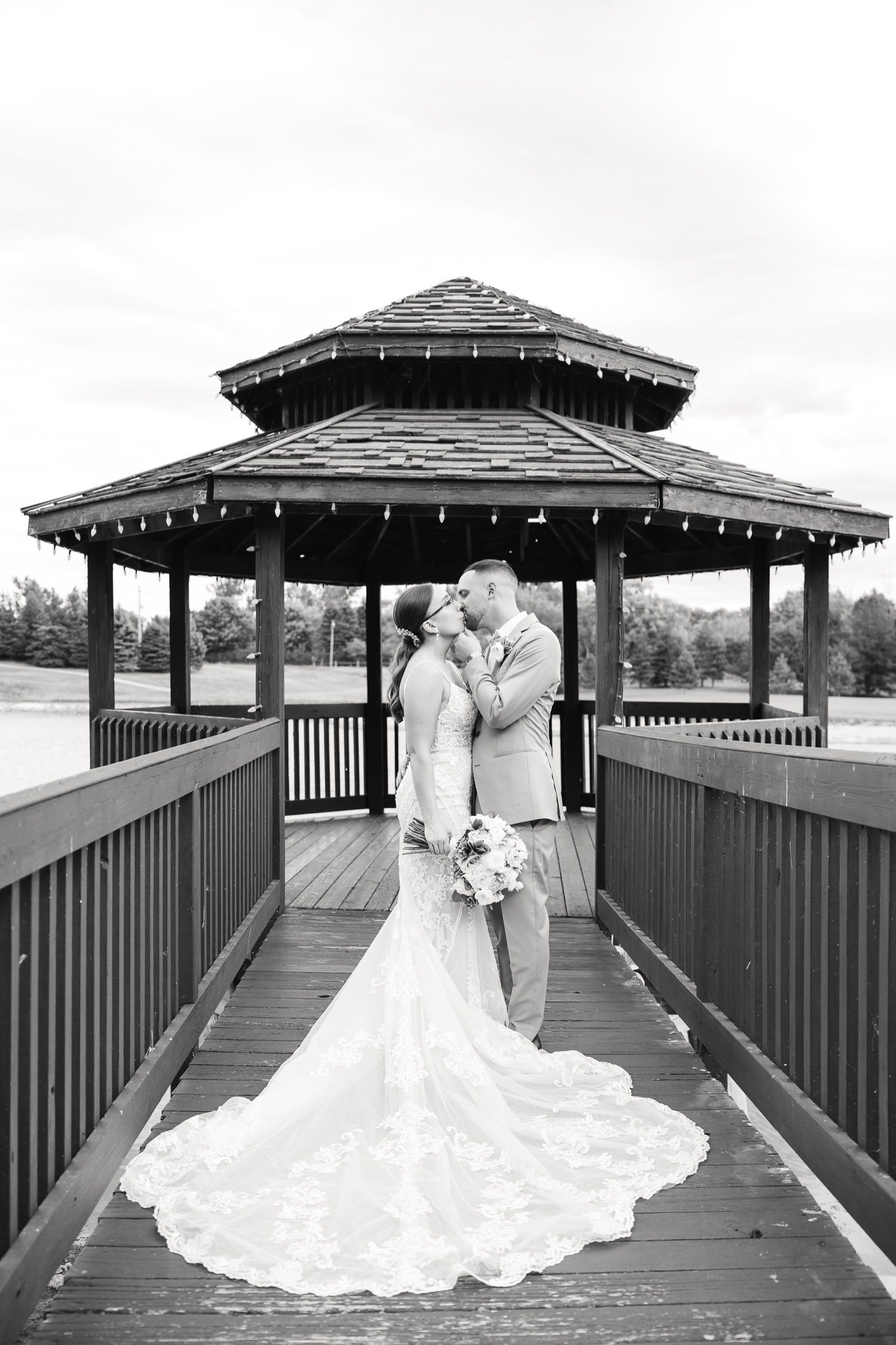 A romantic wedding photo shows a couple embracing under a wooden gazebo with string lights and a flowing lace train.