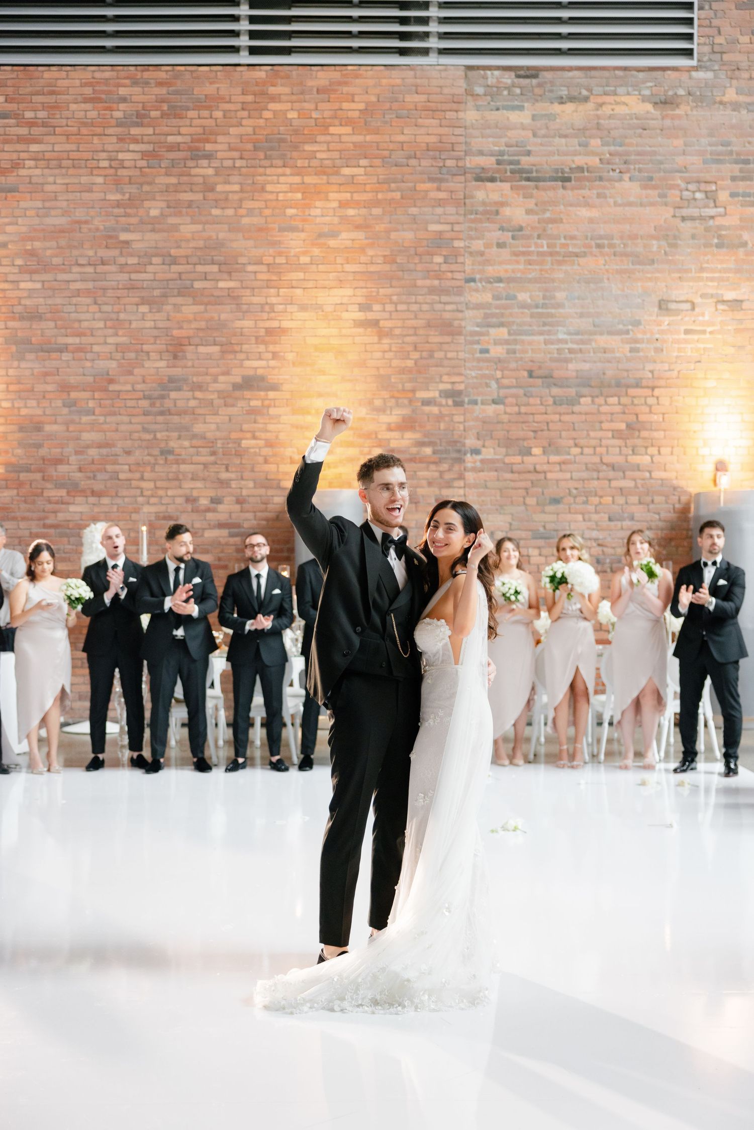 Wedding couple celebrating with wedding party against exposed brick wall in modern white venue.