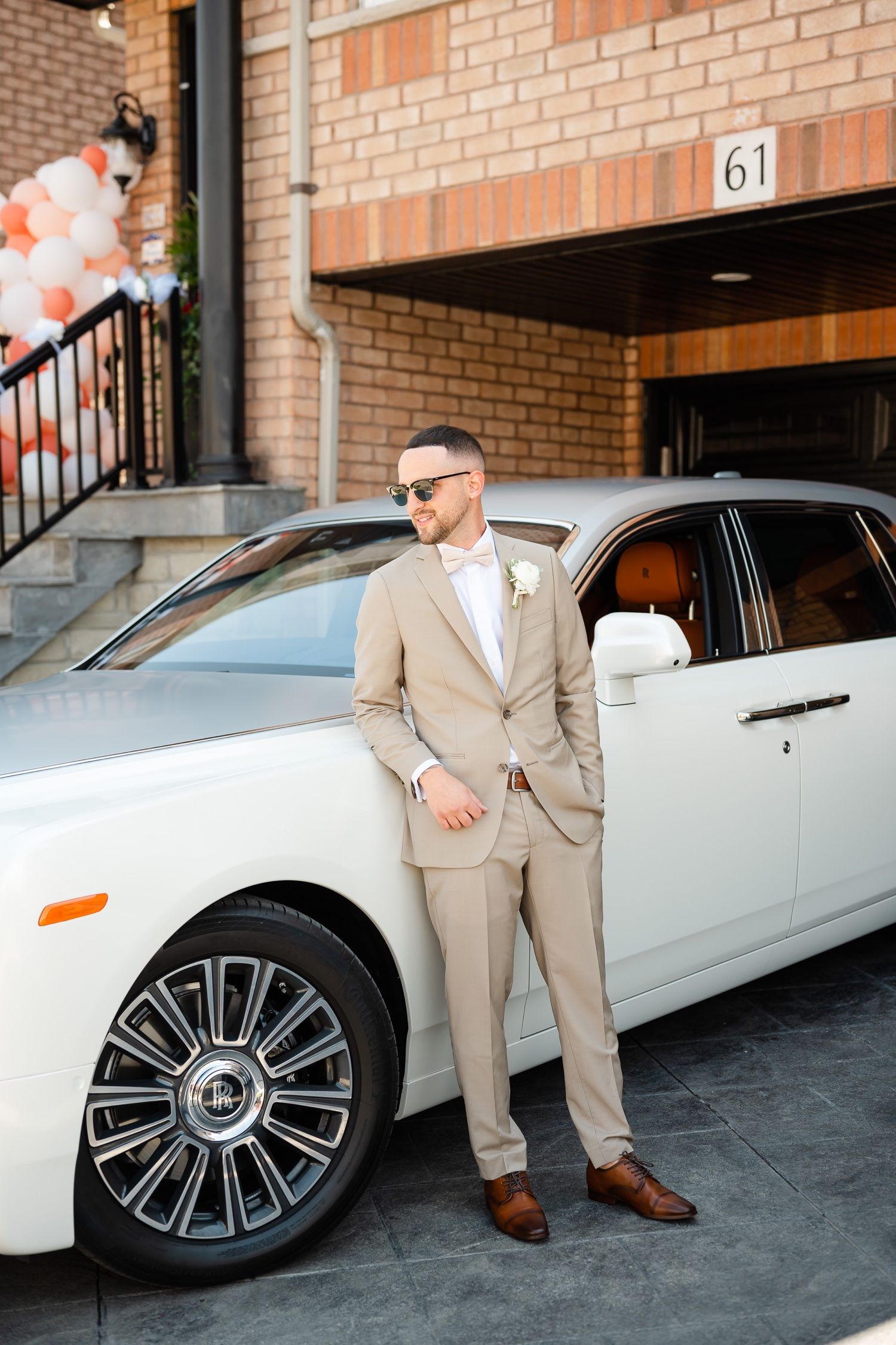 Groom in beige suit poses next to white luxury car outside brick building.