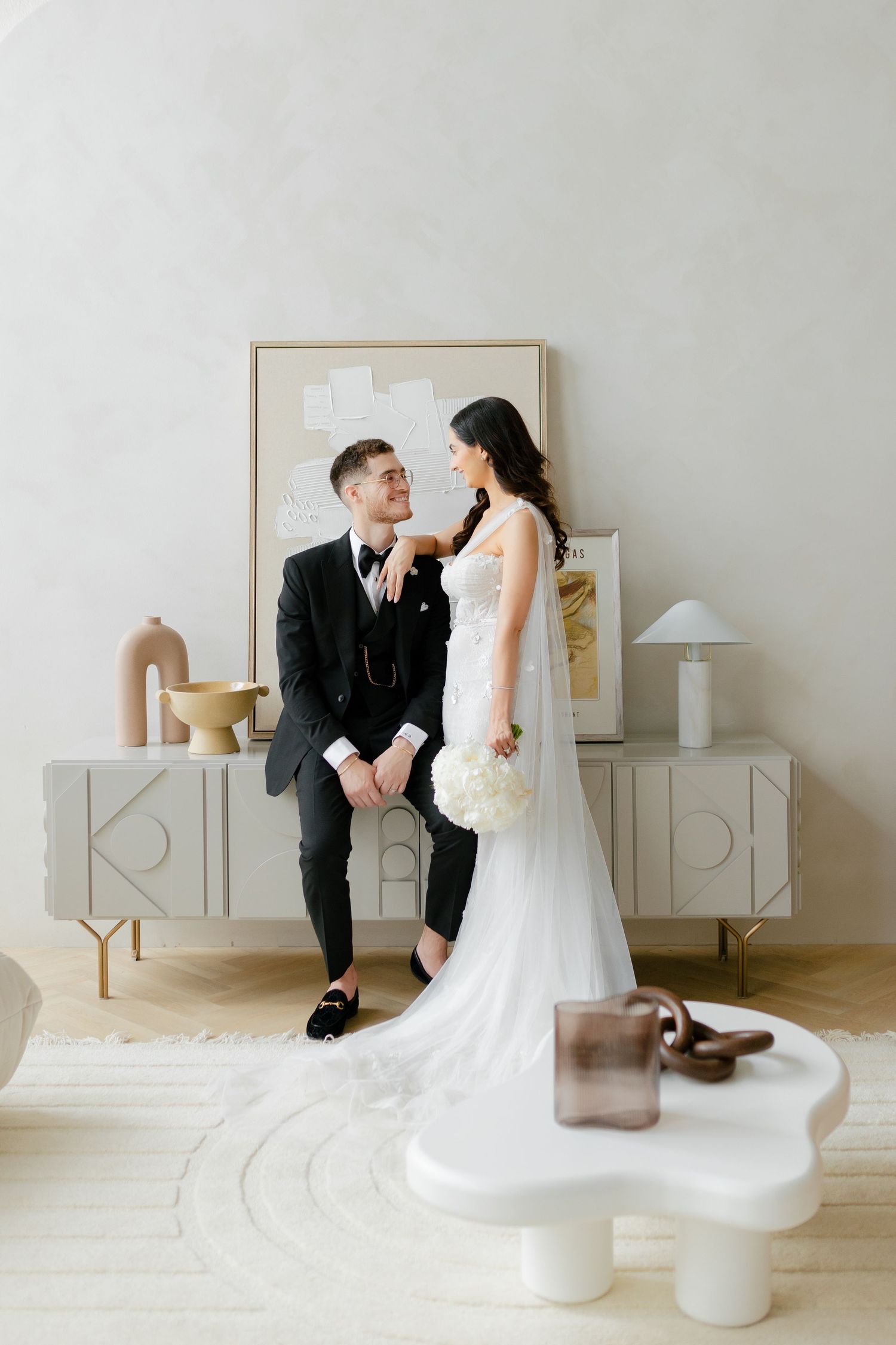 Intimate wedding photo of couple sitting on modern console table in minimalist white room.