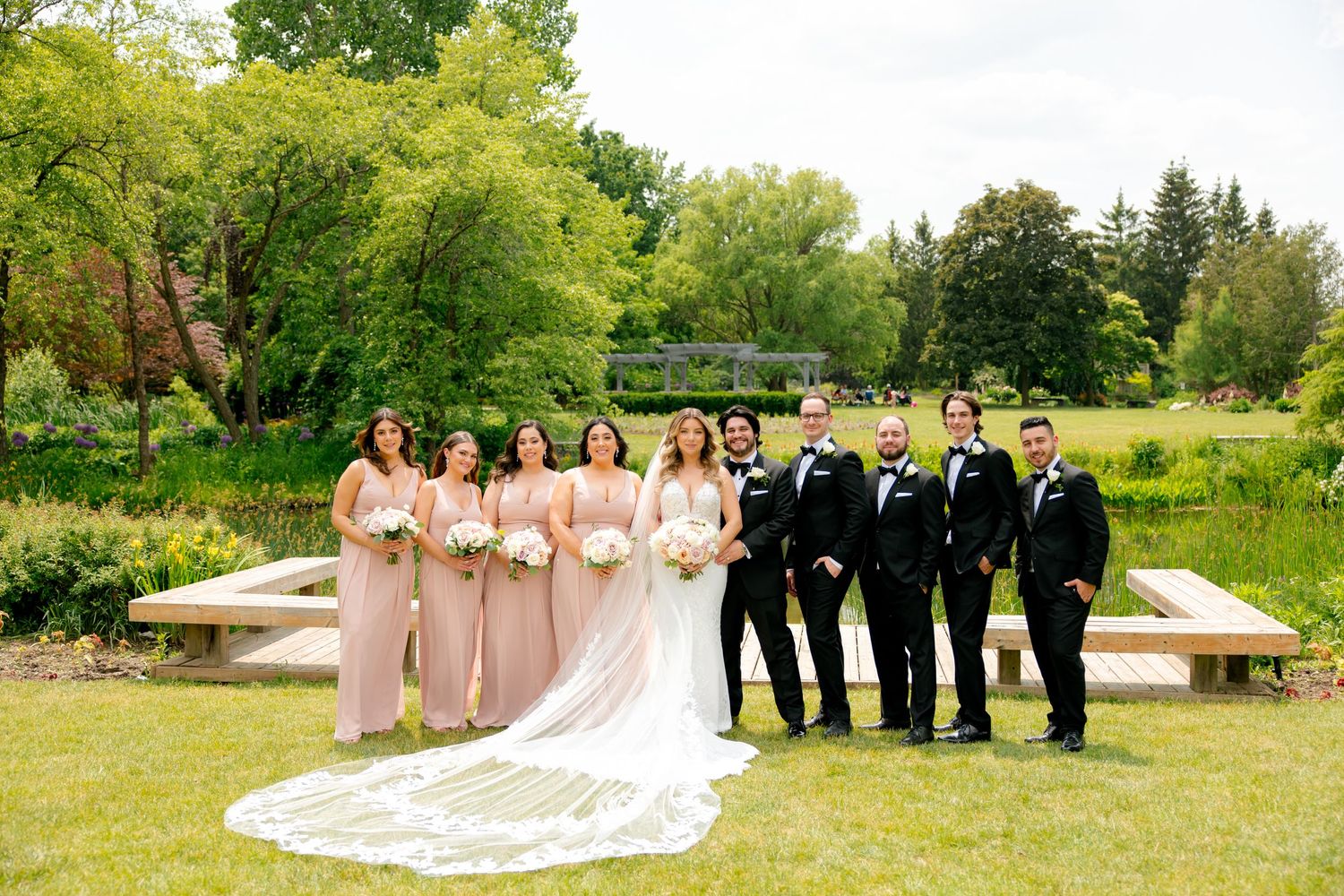 Wedding party poses on wooden boardwalk in lush garden setting with long flowing veil and pink bridesmaids dresses.