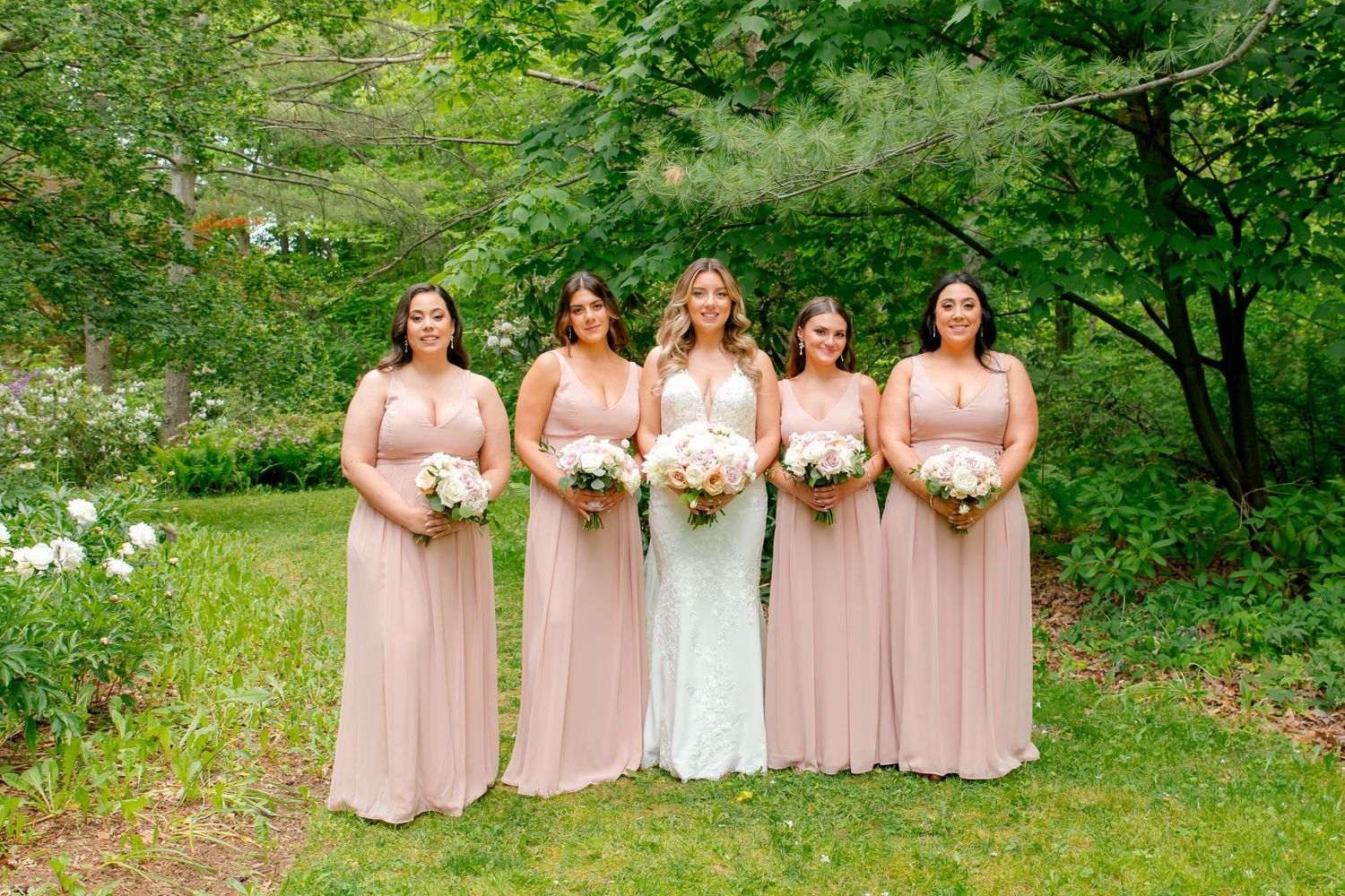 Bridal party in blush pink dresses poses together on garden lawn with white floral bouquets.