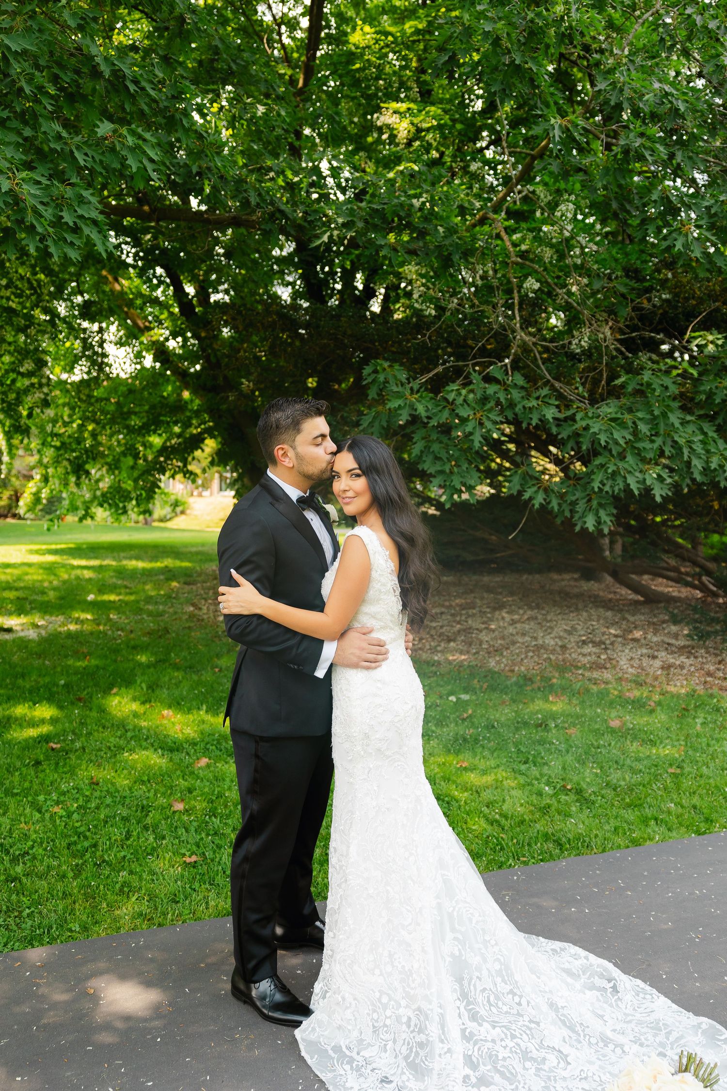 Bride in flowing lace gown and groom in tuxedo embrace under green trees in a garden setting.