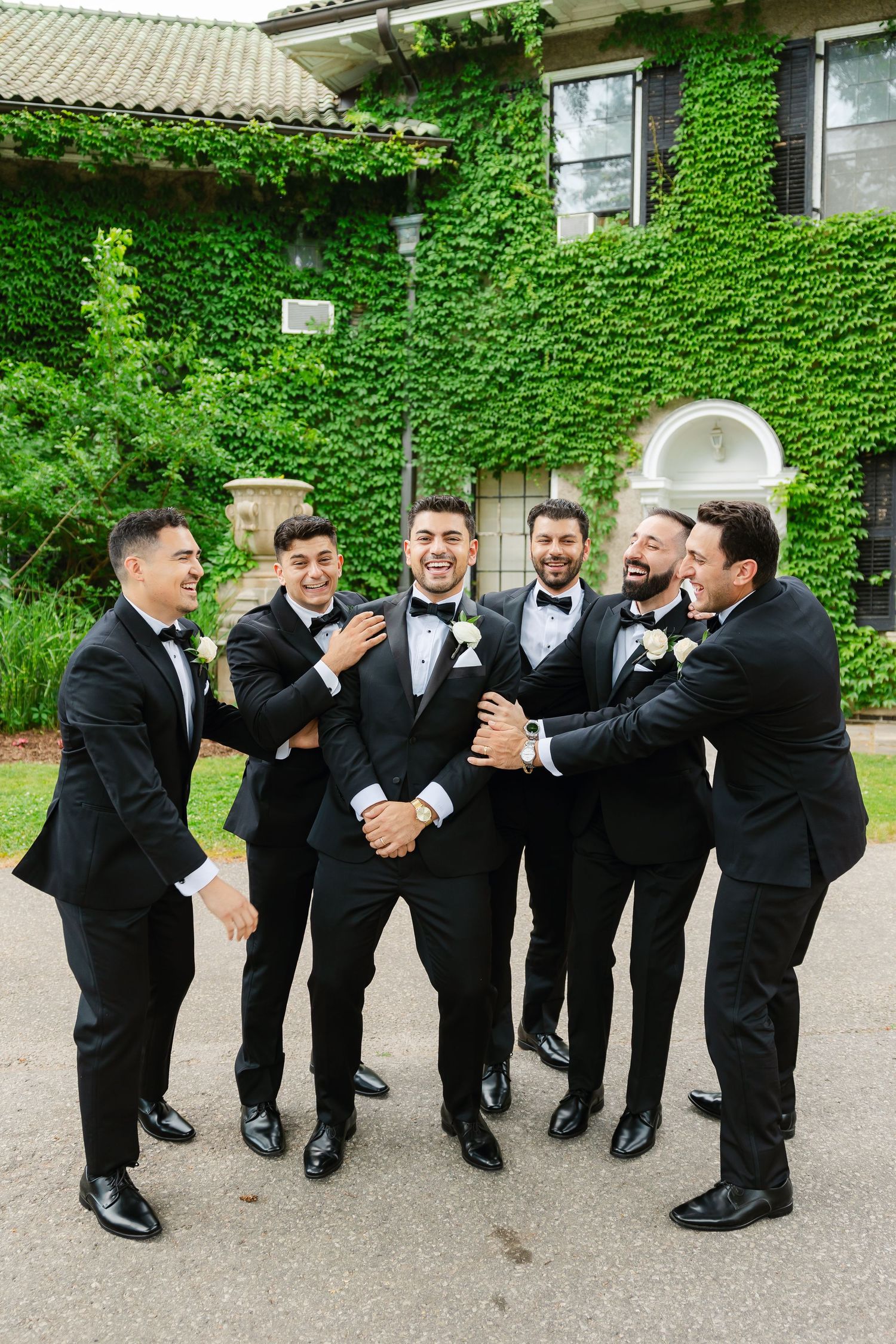 Groomsmen share joyful moment in formal attire against ivy-covered building backdrop.