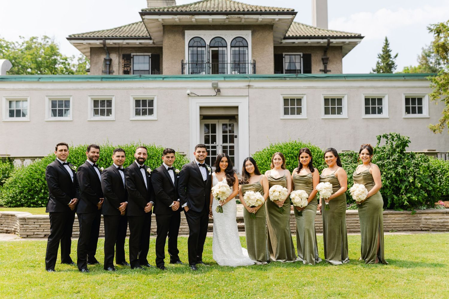Wedding party in sage green dresses and black tuxedos pose outside elegant white mansion.