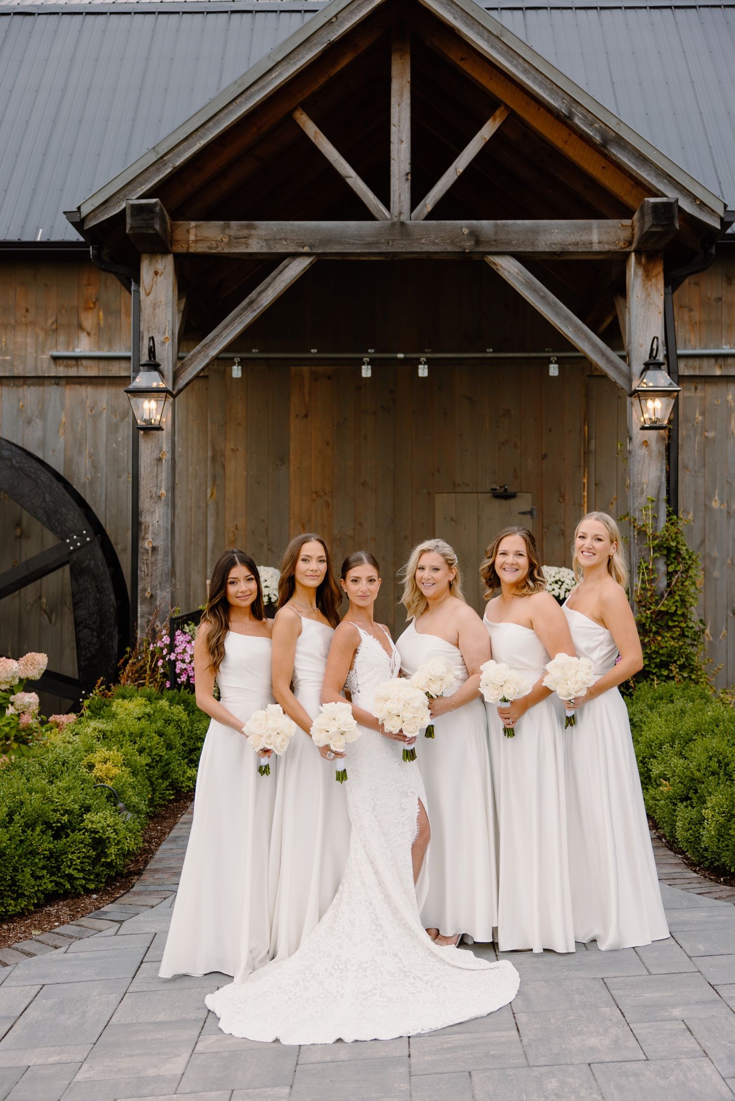 Wedding party poses under a rustic wooden pavilion with string lights and floral landscaping.