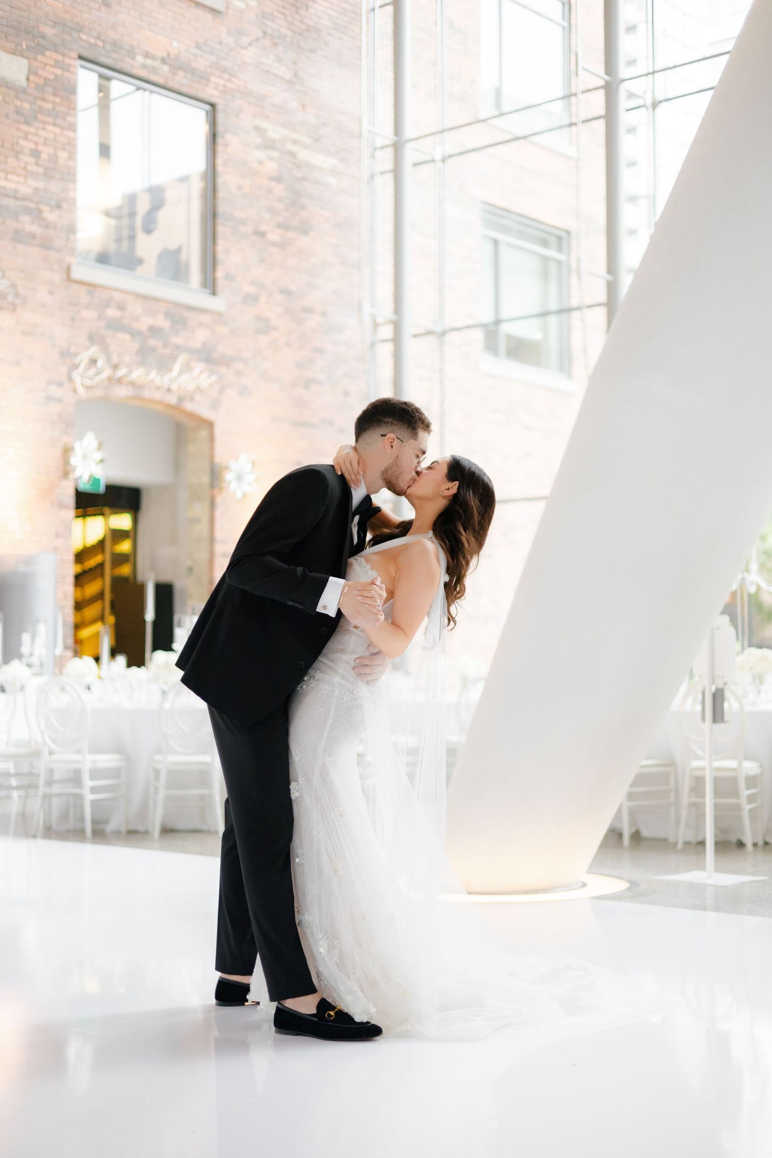 Romantic wedding couple sharing a kiss in an elegant white reception venue with exposed brick walls.