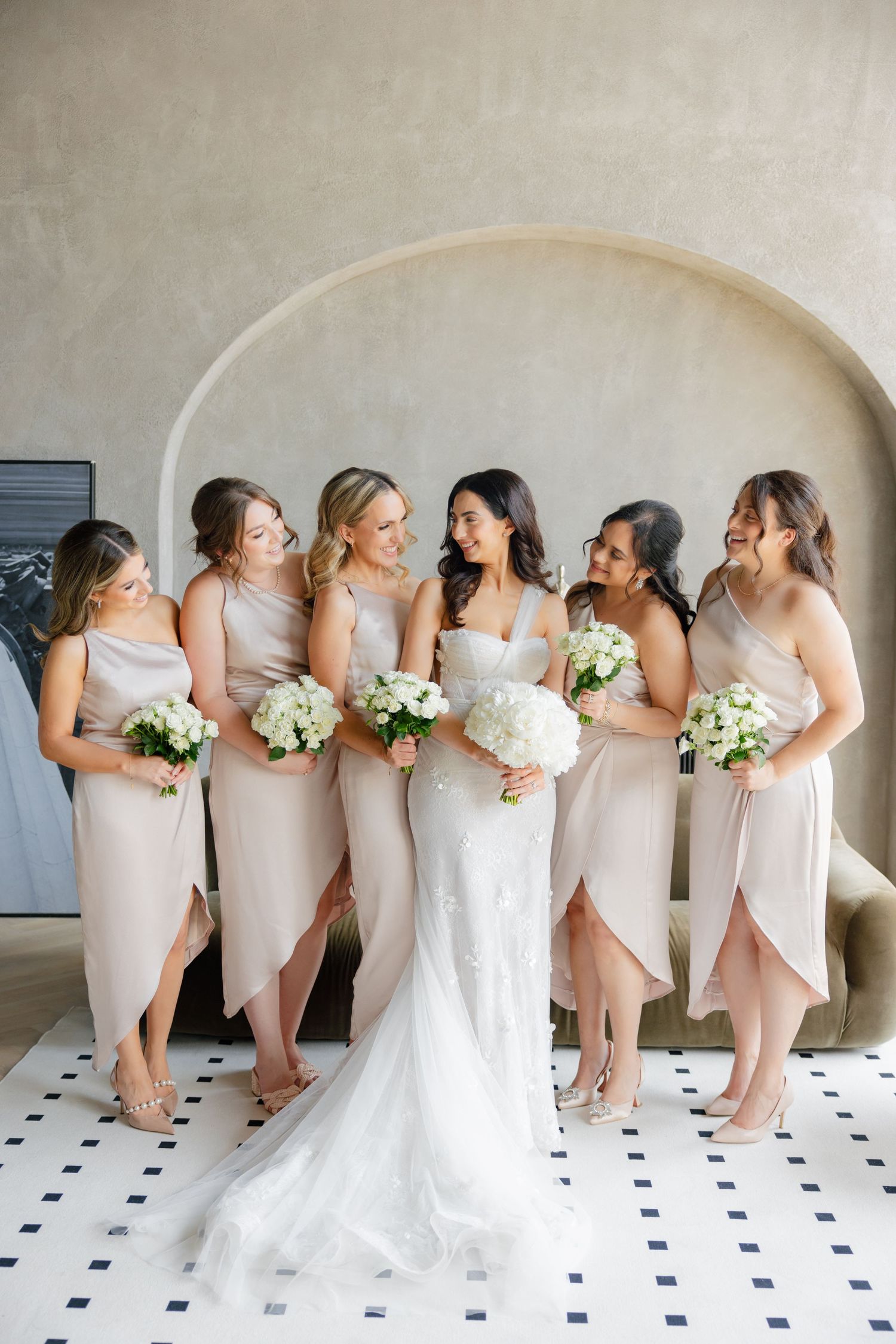 Bridal party in pale pink bridesmaid dresses holding white bouquets pose together in an elegant indoor setting.