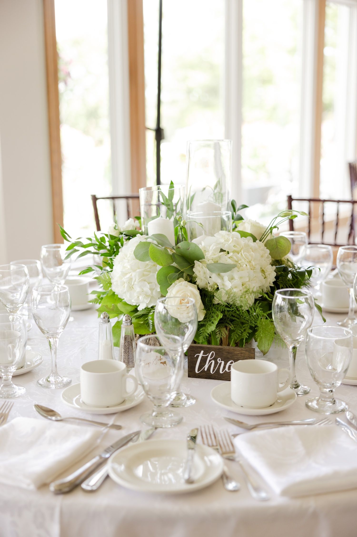 White hydrangea and rose centerpiece with green foliage on round table with elegant place settings.