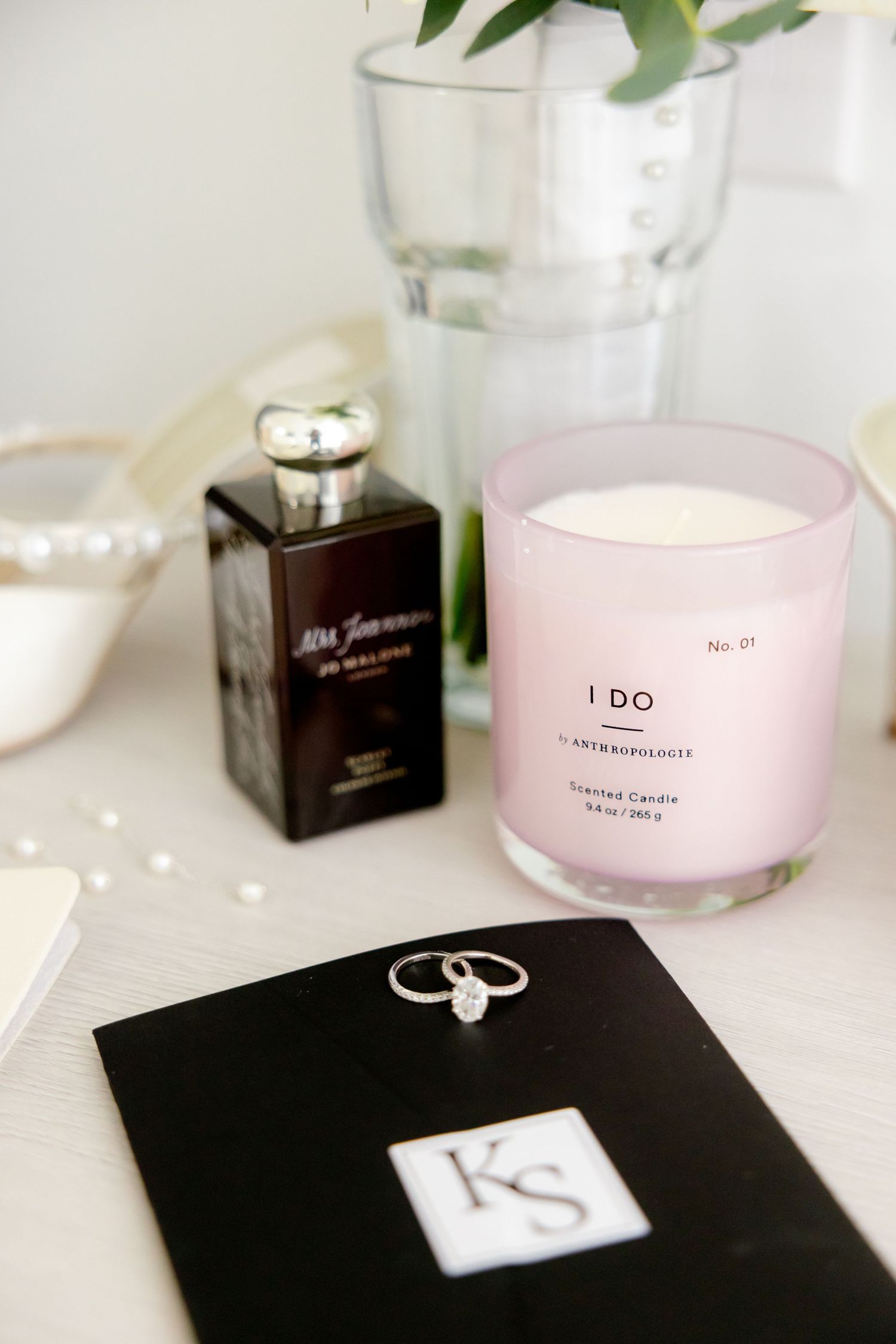 Wedding details with pink candle, black notebook, perfume bottle and engagement ring on white surface.