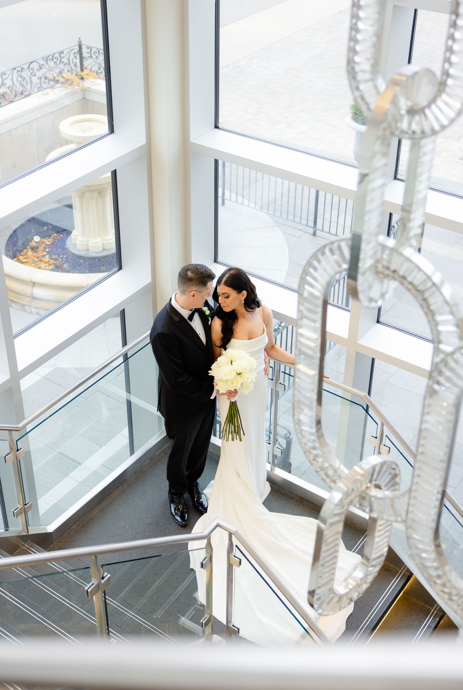 Overhead shot of newlyweds embracing on a modern staircase with glass walls and decorative lighting.