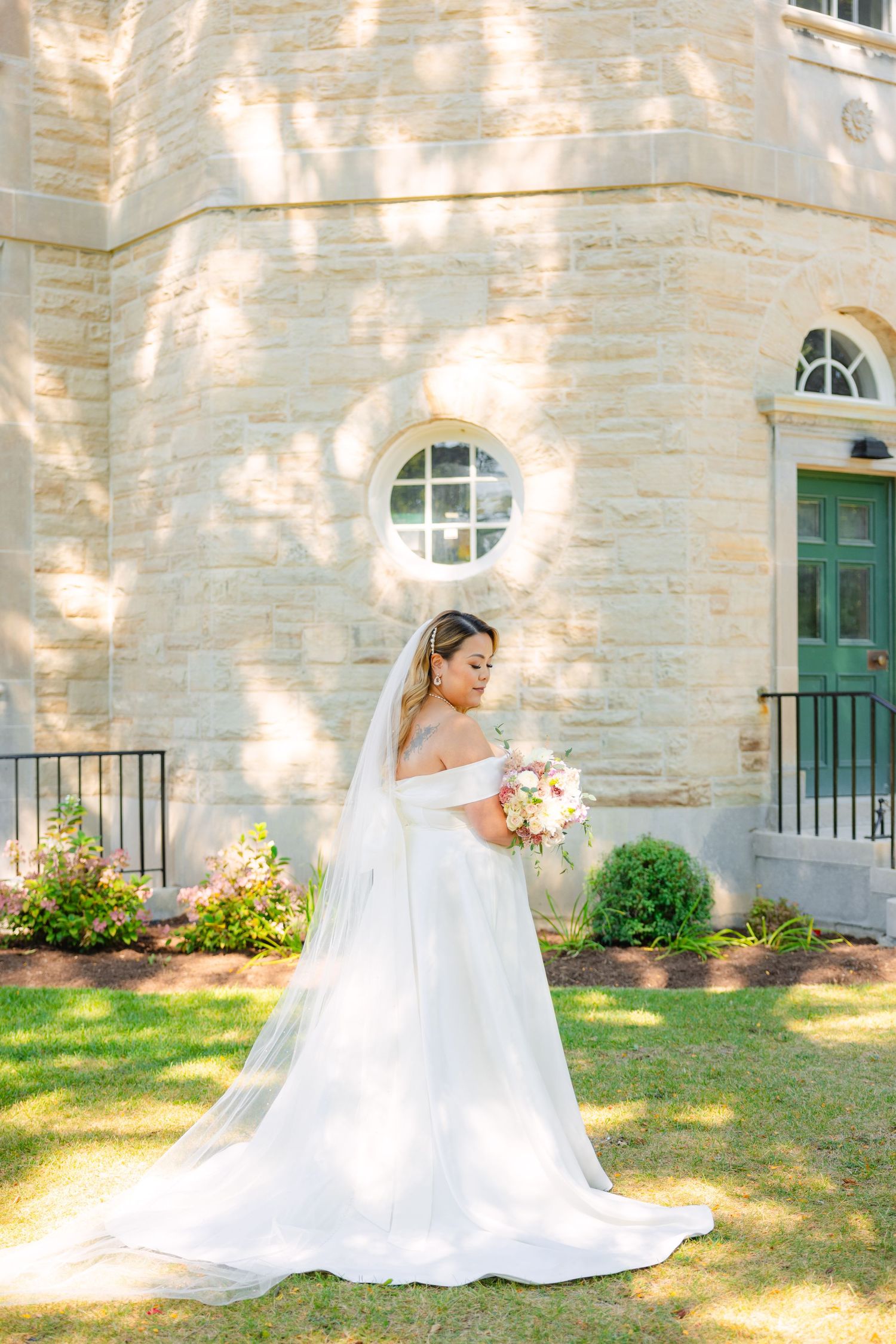 Elegant bride in off-shoulder wedding dress holds bouquet while posing in front of historic stone building.