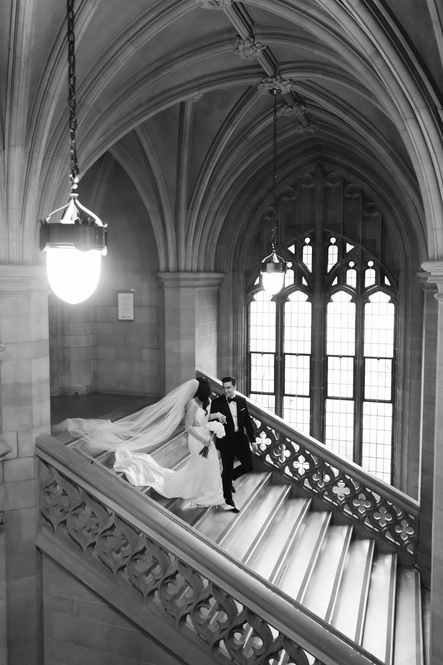 Black and white photo of newlyweds on elegant stone staircase with gothic architectural details.
