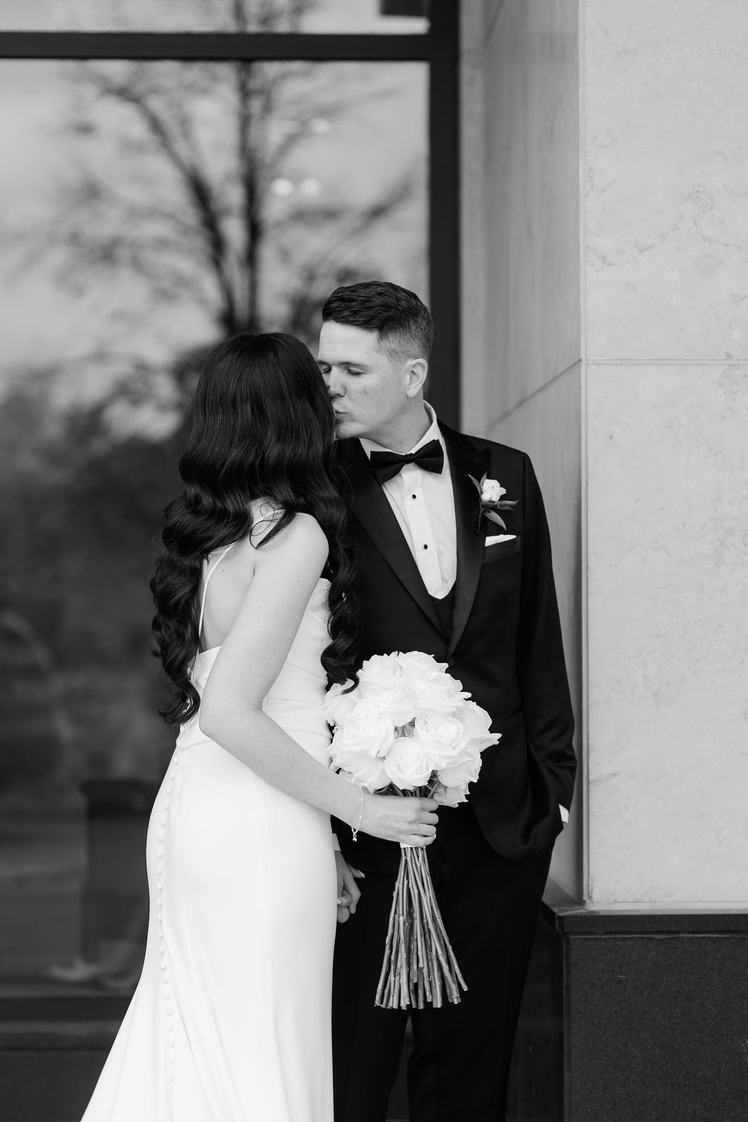 A bride holding white flowers poses with her groom in formal attire against architectural columns.